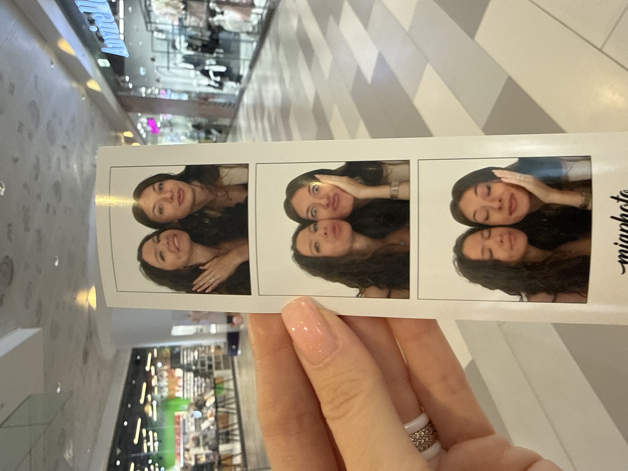 The image shows a strip of photo booth photos being held up by a person's hand. The photo strip features three pairs of photos, each of two young women. In the first panel, the women are smiling and looking at the camera. The woman on the left has her hand on her chin. In the second panel, both women are making exaggerated facial expressions, with one woman puckering her lips towards the other, who has her hand on her cheek. In the third panel, both women have their eyes closed and are smiling, with the woman on the right touching her cheek. The background of the photo booth images appears to be a plain white backdrop.
The person holding the photo strip has painted fingernails and is wearing a ring with small stones. The background of the overall image shows a well-lit indoor shopping mall with stores visible, including one with a sign that appears to say "Anniel". The floor of the mall has a patterned tile design. The lighting in the mall appears to be artificial, with overhead lights visible. The scene suggests a casual outing in a public shopping area. Without additional context or verifiable landmarks within the image, it is not possible to definitively determine the specific location in Moscow, Russia.