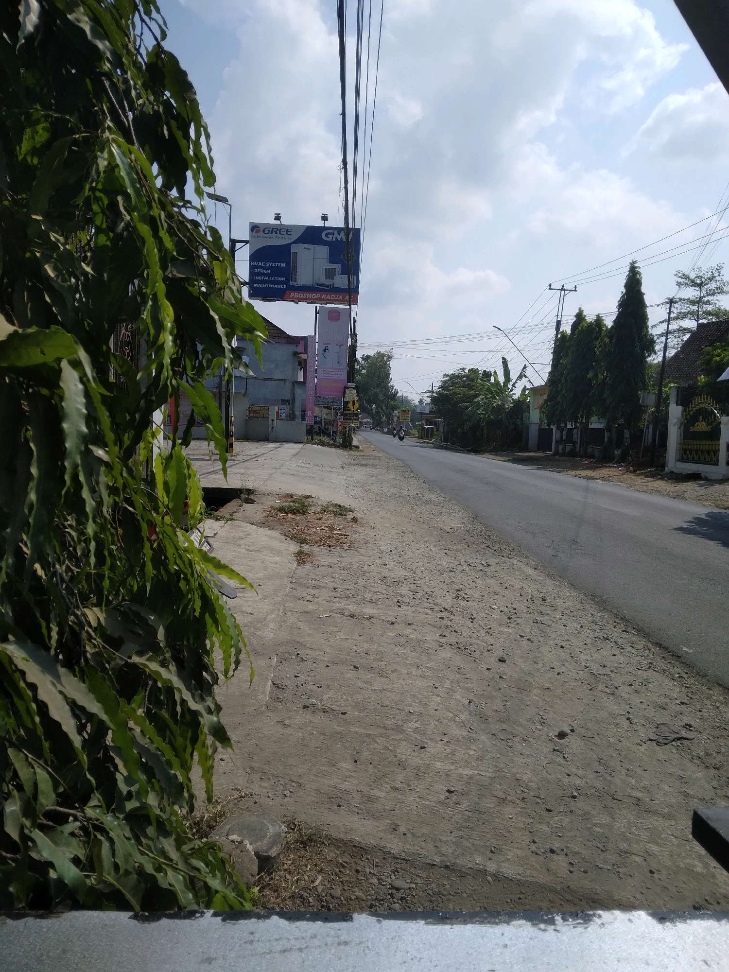 The image captures a street scene in Sokaraja, Indonesia, under a bright, cloudy sky. In the foreground on the left, lush green foliage hangs down, partially obscuring the view. To the right of the foliage is a concrete sidewalk, transitioning to a gravelly area bordering a paved road. The road stretches into the distance, with a motorcycle visible in the middle ground.
Along the side of the road, several buildings are present. A prominent billboard features the GREE brand and details about HVAC systems, along with smaller advertisements below it. Further down the street, trees line the roadside, and houses with traditional Indonesian architecture are visible, some featuring decorative fences. Utility poles and power lines are strung across the sky, a common sight in urban and semi-urban areas. The overall impression is that of a sunny day in a moderately developed area.