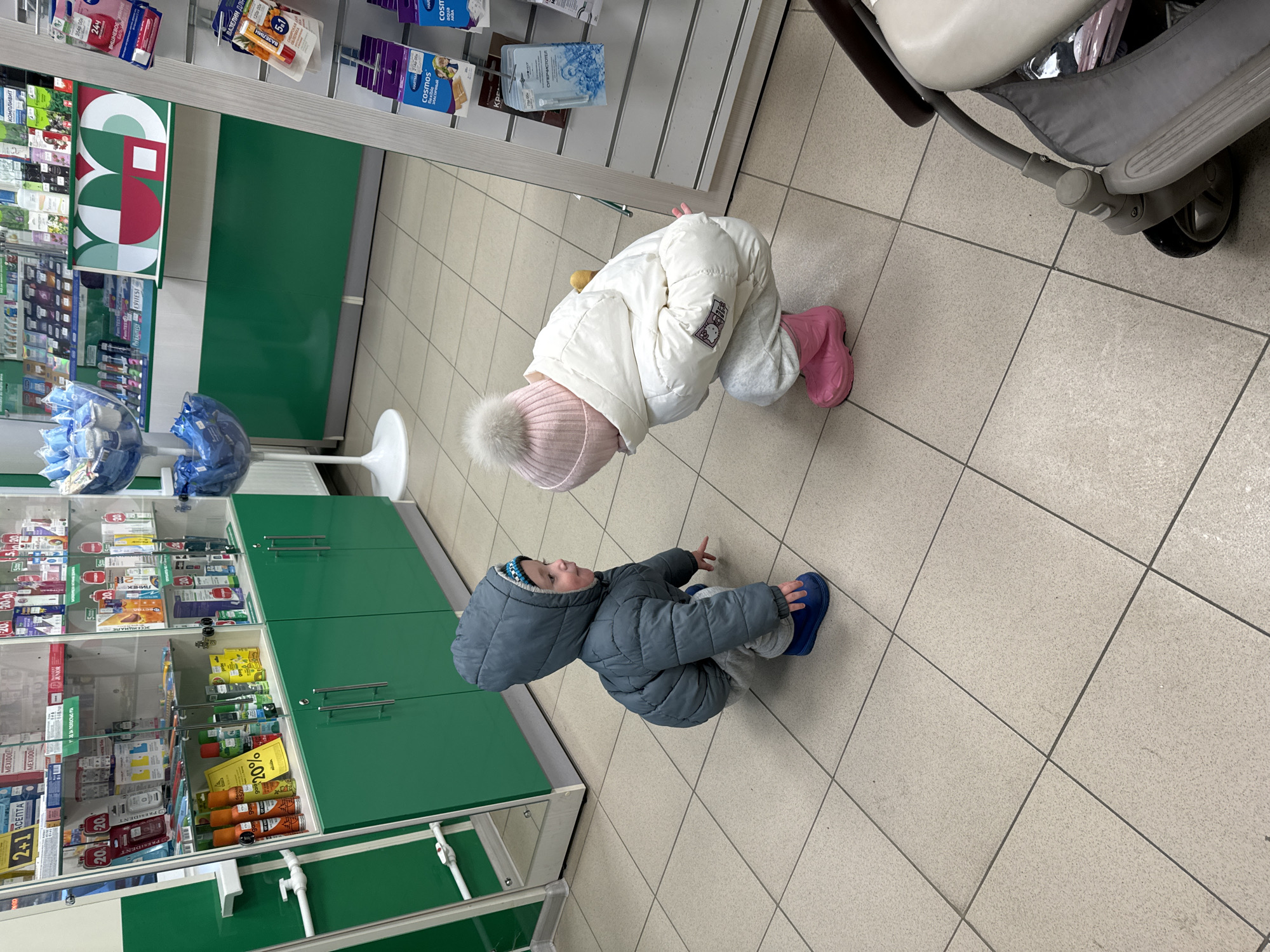 The photograph shows two young children inside a pharmacy or store that likely sells health and personal care products. The setting is likely Odintsovo, Russia based on visible product packaging on shelves.
One child, wearing a white puffy jacket, light gray pants, and pink boots and hat, is bending forward, looking down at something. The other child, dressed in a gray puffy jacket, light gray pants, and blue boots, is sitting on the floor, looking upward.
The environment includes shelves stocked with medications, personal care items, and other products. The shelves are green and white, and a stroller can be seen in the upper right corner of the photo. The floor is tiled.