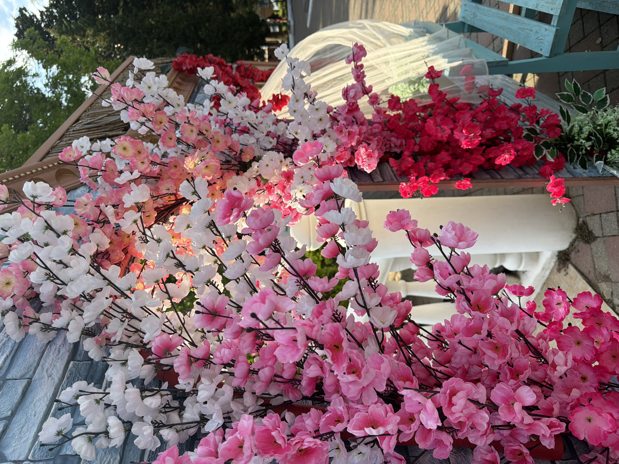 An outdoor scene features artificial floral arrangements, predominantly pink and white cherry blossoms, cascading down the side of a decorative structure. The structure appears to be a whimsical, small building with a wooden roof and a textured wall resembling blue bricks. Behind the flowers, a white, sheer fabric drapes over an unseen object, possibly part of the decorative setup. To the right, a section of a light blue wooden bench is visible. The ground is paved with bricks. In the background, trees with green foliage and a glimpse of a road suggest an urban or park setting. The lighting indicates it is daytime. The image is a close-up view focusing on the colorful flowers and decorative elements, with a shallow depth of field that blurs the background. The location is not specified within the image itself.