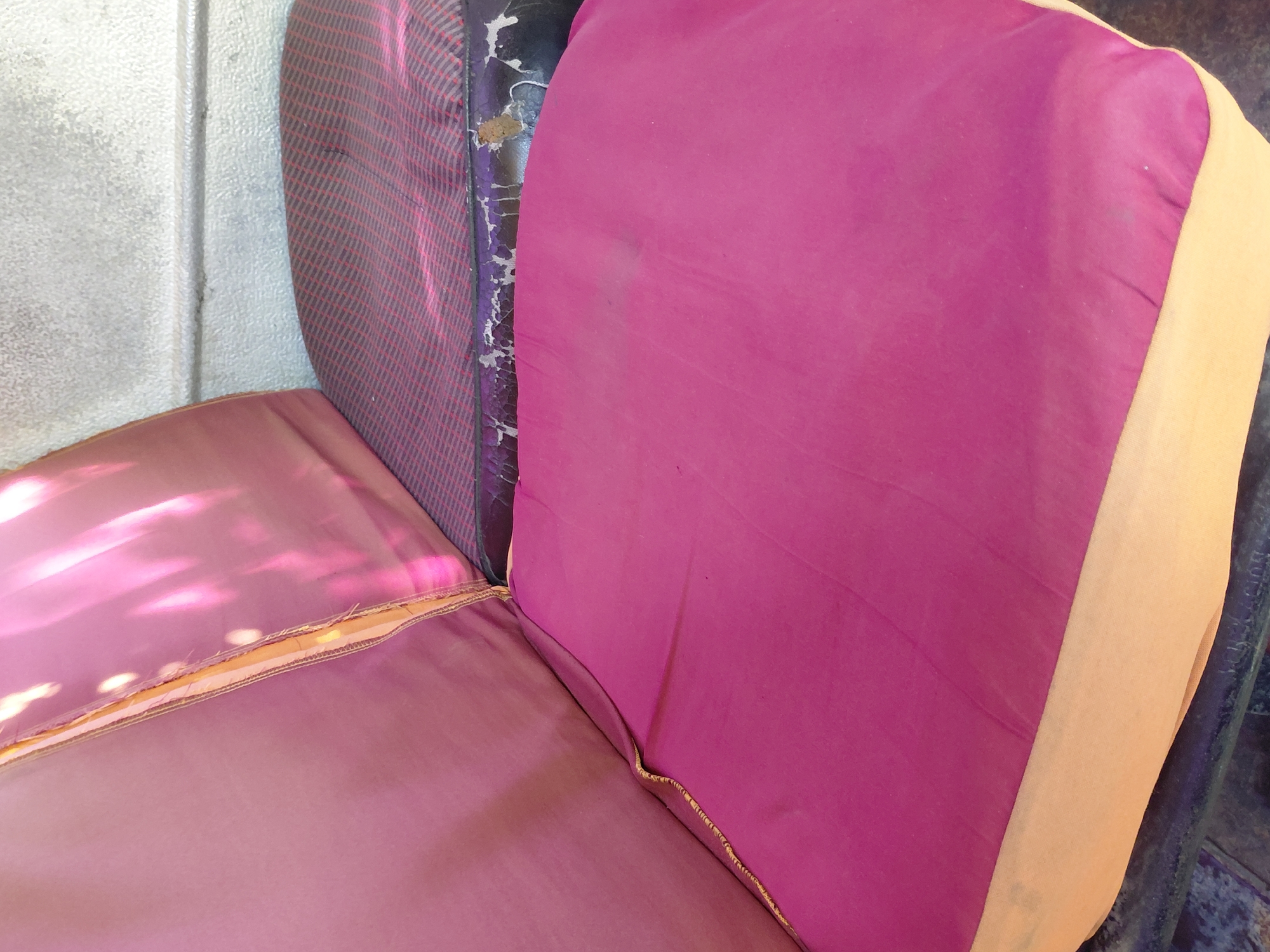 The image shows a close-up view of worn, padded seating, likely from an outdoor bench or public seating area. The seating is upholstered in a faded pink fabric with a light beige underside. There is visible damage to the fabric, including tears and a dark, worn section. The background appears to be a textured, light gray wall. The lighting suggests an outdoor environment, possibly under sunlight, as indicated by the bright reflections on the pink fabric. The scene is set in Santa Ana, Venezuela.