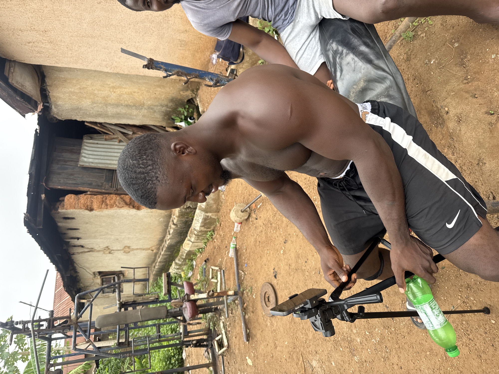 An individual with dark skin, a muscular build, and short, dark hair is seated outdoors, facing right and slightly away from the camera. They are wearing black athletic shorts with a white stripe. Their bare chest is visible, revealing a scar on the upper right arm. They are holding a green plastic bottle in their left hand and their right hand is on a tripod that holds a smartphone. The ground is dirt, with scattered debris including weight plates and bottles. Behind the individual, there is a makeshift outdoor gym setup with metal equipment. Further in the background, there is a simple building with a corrugated metal roof and mud walls. Another individual with dark skin and short hair, wearing a gray t-shirt and grey shorts, is partially visible on the right side of the frame, seated next to the primary subject. The overall scene appears to be in Akwanga, Nigeria.