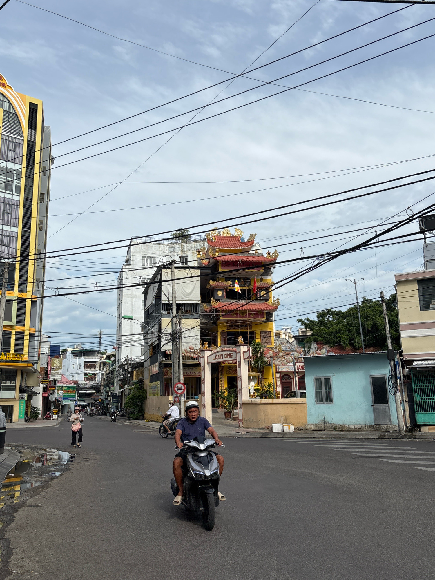 The image depicts a street scene in Nha Trang, Vietnam, on an overcast day. In the foreground, a man wearing a dark blue t-shirt, white helmet, and light-colored shorts rides a black scooter. His feet are clad in light-colored slip-on sandals. To the left, a woman in a light-colored top and pants, with a dark bag slung over her shoulder and a hat, walks on the sidewalk. She is looking towards the left of the frame.
The street is paved with asphalt and shows signs of recent rain, with puddles reflecting the surrounding buildings and sky. Overhead, a dense network of power lines crisscrosses the frame.
Dominating the midground is a multi-tiered building with traditional Vietnamese architecture, featuring red tiled roofs with ornate golden trim. The building is painted yellow and appears to be a temple or pagoda. Text on the building reads "LÀNG ONG". To the right of this structure, a small, light blue building with a single window and a door is visible. Further to the right, a building with a green metal gate is present.
In the background, a tall, modern building with a yellow facade and arched windows stands prominently on the left. Other buildings of varying heights and architectural styles line the street, creating a dense urban environment. Trees are visible in the distance, peeking over the rooftops. The sky is filled with clouds, suggesting a potentially humid or rainy climate. The overall impression is a busy, bustling city street with a blend of modern and traditional elements.
