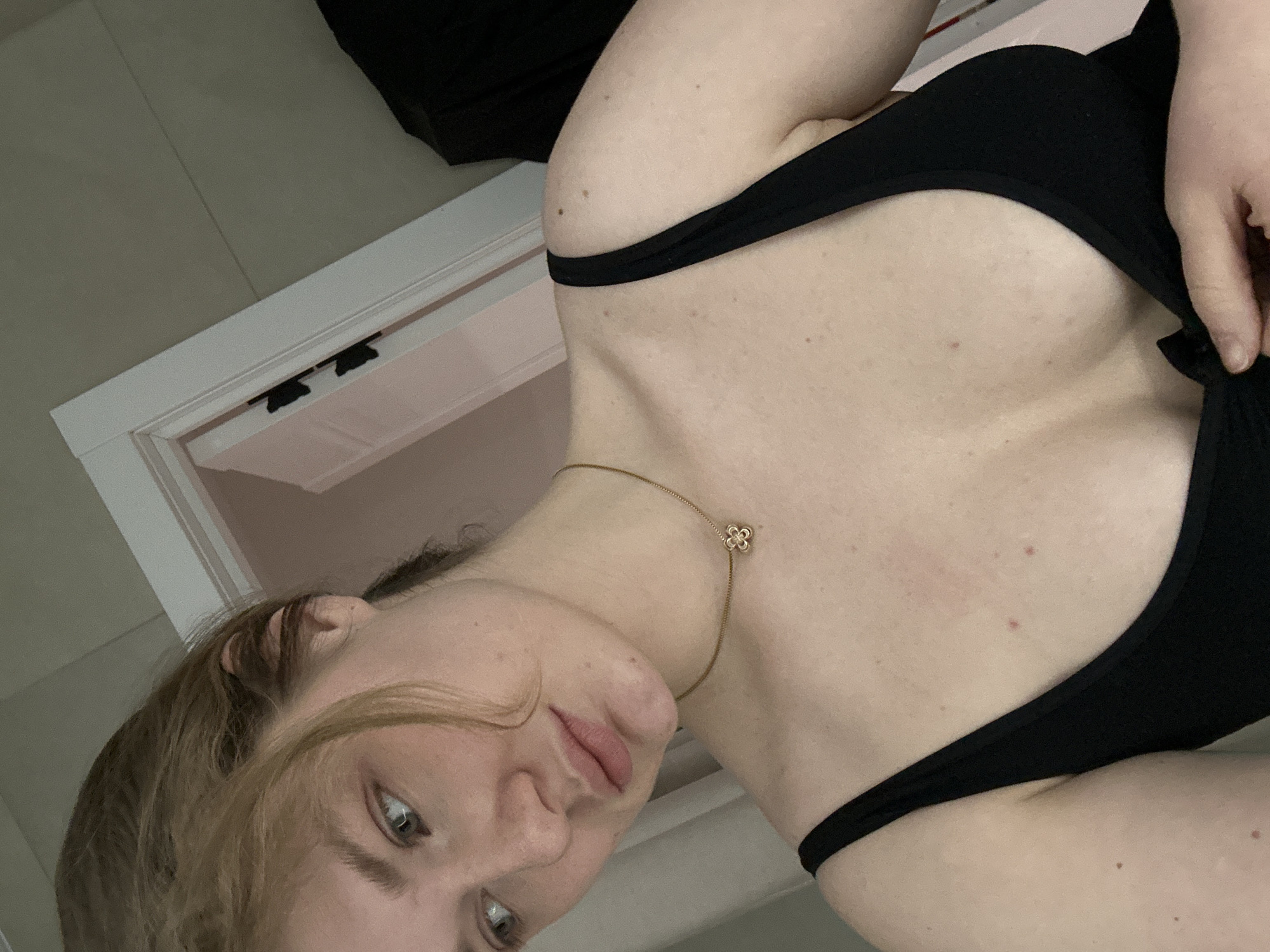 A selfie taken indoors, with the subject, a young woman, facing downward and slightly to the side. She has fair skin, blonde hair, and is wearing a black top with thin straps. A delicate gold necklace with a clover-shaped pendant is visible around her neck. The background includes a white doorframe and a glimpse of what appears to be a white wall and a dark object. The environment is indoors, but the specific location in Kazan, Russia, cannot be determined from the image.