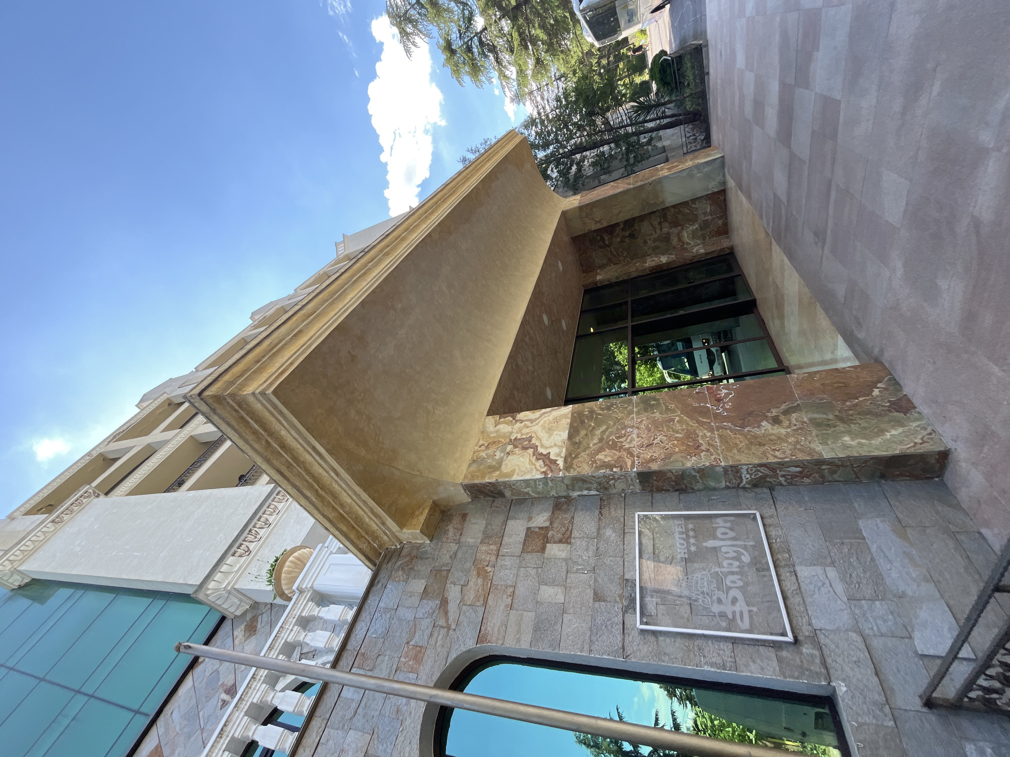 This is a medium-angle, eye-level shot of the exterior of the Hotel Babylon in Nesebar, Bulgaria. The building features a modern architectural style with a facade of stone and marble. An ornate, golden-colored overhang extends from the building, casting a shadow on the entrance below. A sign on the wall reads "HOTEL Babylon" with four stars. The image captures a clear blue sky with some white clouds, suggesting it is daytime, likely sunny. Lush green trees are visible in the background, reflected in the building's glass windows. The overall scene conveys a sense of elegance and tranquility, with the sunlight highlighting the building's luxurious materials.