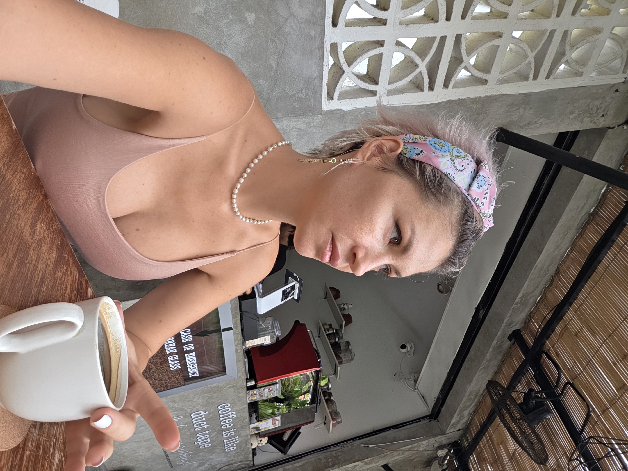 The media file shows a woman with short, pink-tinged hair, wearing a pink camisole and a patterned headband. She is seated at a wooden table, holding a white mug of coffee. A pearl necklace adorns her neck, and a delicate earring dangles from her ear. Her gaze is directed downwards, with a thoughtful expression.
The setting appears to be an outdoor cafe, possibly in North Kuta, Indonesia, given the humid climate implied by the presence of a ceiling fan and bamboo blinds. The background reveals a concrete wall with decorative lattice work, a coffee machine, shelves stocked with jars, and what looks like a framed picture. Visible text on a framed item reads: "IN CASE OF EMERGENCY BREAK GLASS" and "coffee is like duct tape...it fixes everything." The lighting suggests it is daytime, likely morning or afternoon.
