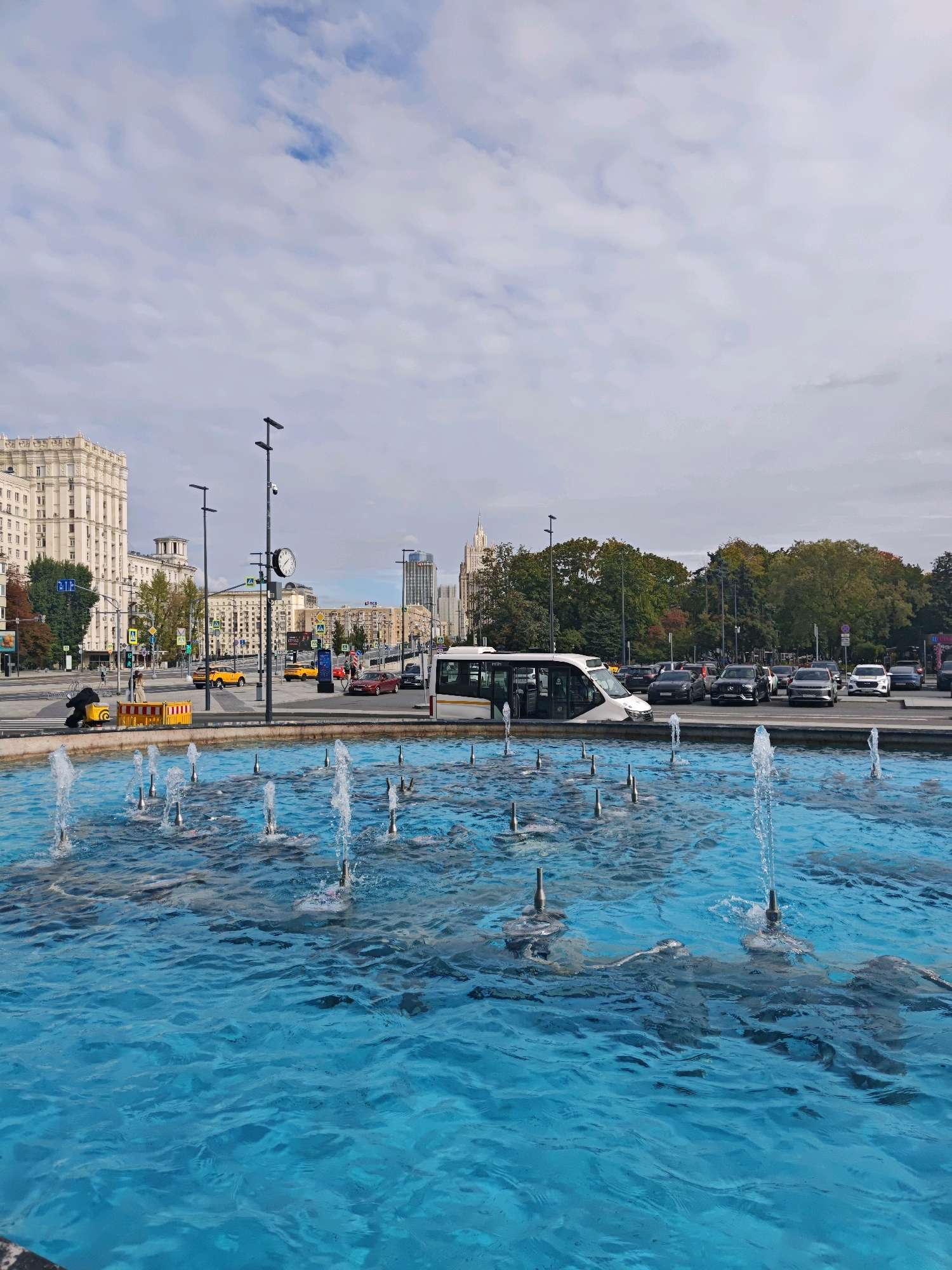 In Moscow, Russia, a public fountain with bright blue water and multiple active jets dominates the foreground. Beyond the fountain, a street scene unfolds under a cloudy sky. A large, light-colored building with a classical facade stands prominently on the left, suggesting a monumental architectural style typical of Stalinist skyscrapers. Traffic moves along the street, with cars of various colors and a white bus visible. Streetlights and traffic signals are present. Trees with green foliage line the background, interspersed with other buildings, including some modern high-rises. The overall impression is of an urban environment, likely a city square or thoroughfare in Moscow, with the fountain serving as a central decorative element.