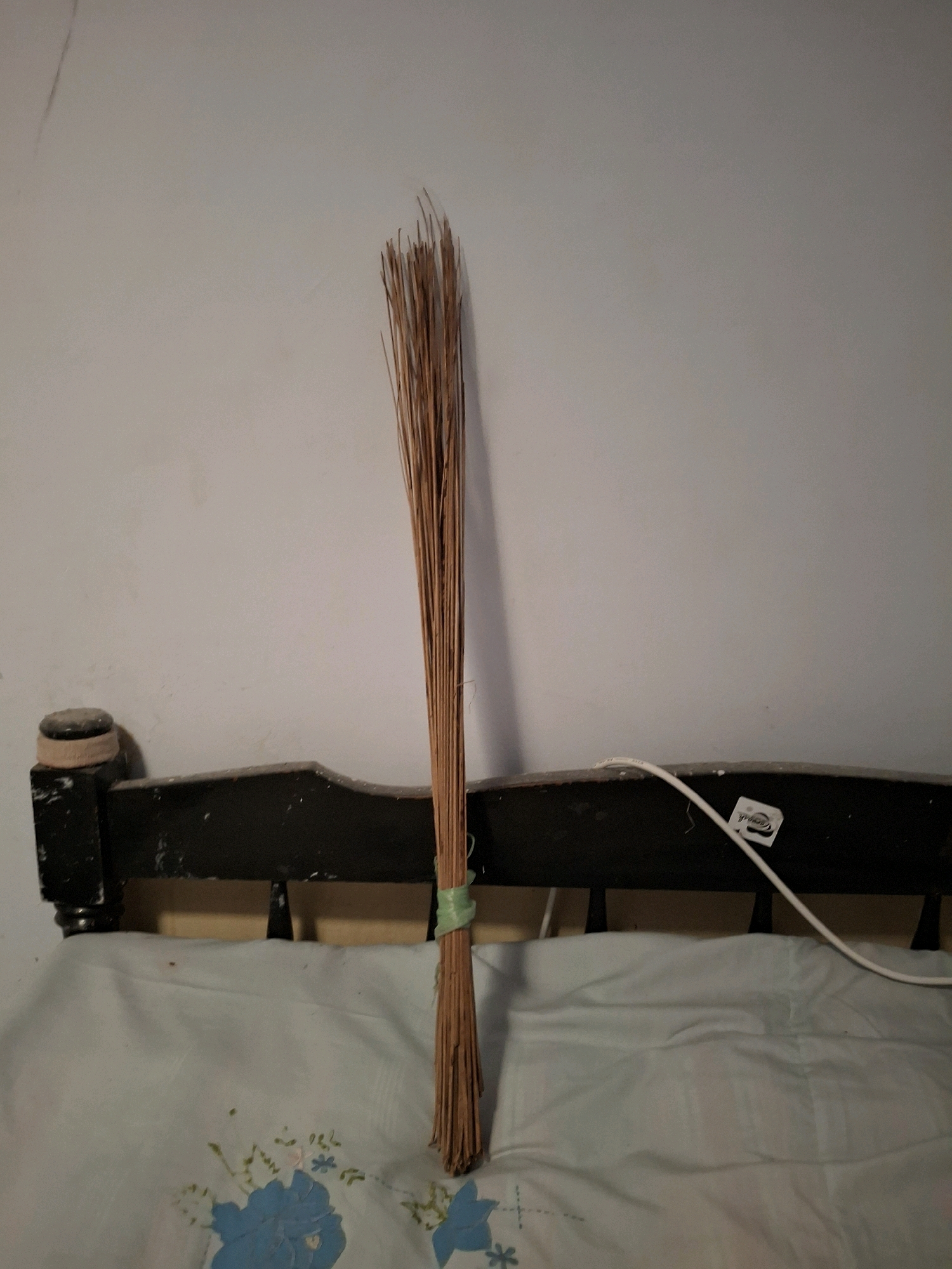 A broom made of dry palm fronds is leaning against the headboard of a bed in Lumajang, Indonesia. The broom has a green band around its middle. The headboard is black with some wear and tear, and it has a rounded, lighter-colored finial on the left side. The bed is covered with a light blue patterned sheet featuring blue and light pink floral designs. A white electrical cord runs across the headboard and onto the sheet. The wall behind the bed is a plain, light gray color.