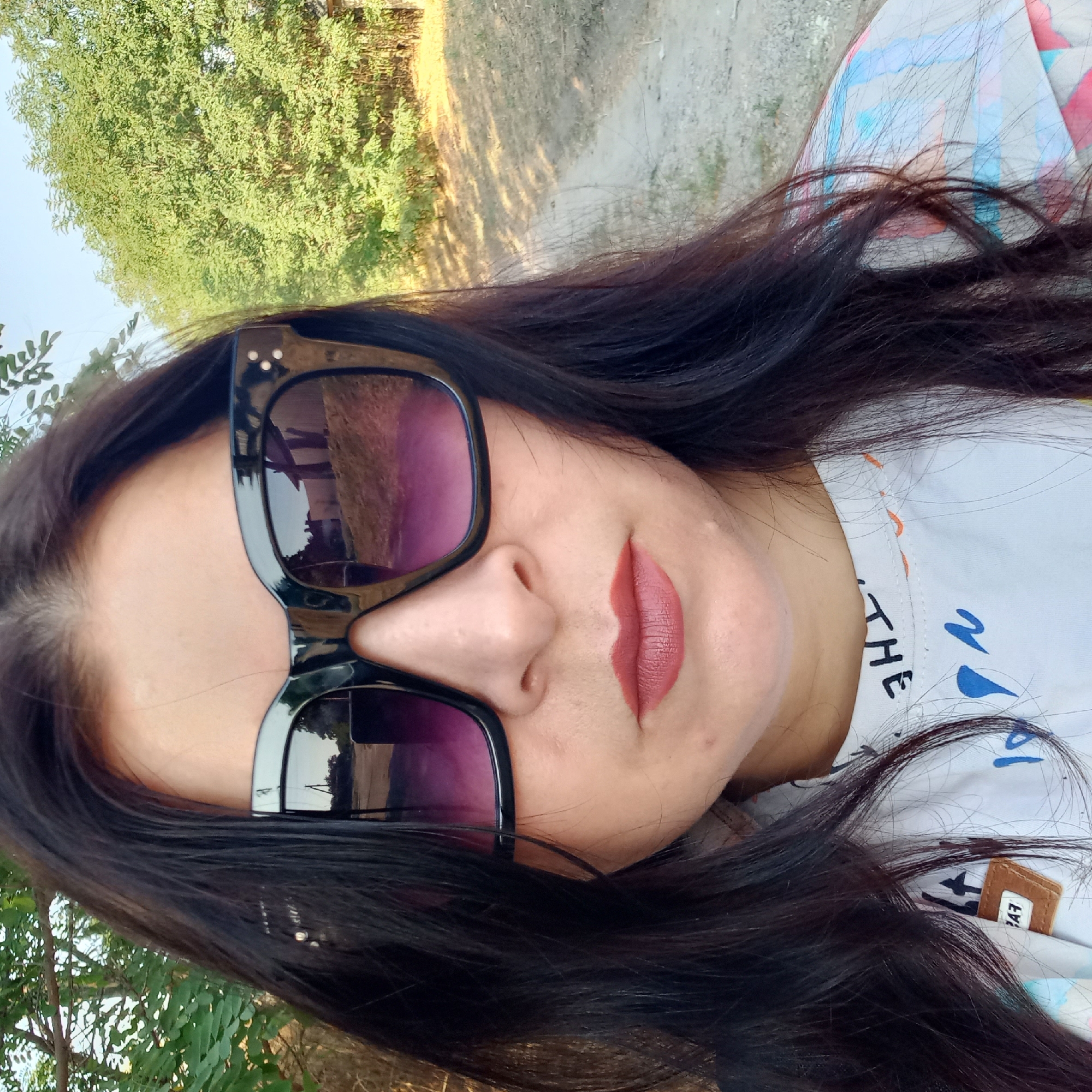 A close-up selfie shows a woman with long, dark hair and large black sunglasses. She is wearing a white shirt with a colorful abstract print. The background appears to be a rural outdoor setting with trees and a dirt path. The woman's mouth is slightly open and she is wearing lipstick. The sunglasses reflect a scene that includes buildings and trees. The image was taken in Klyntsi, Ukraine.