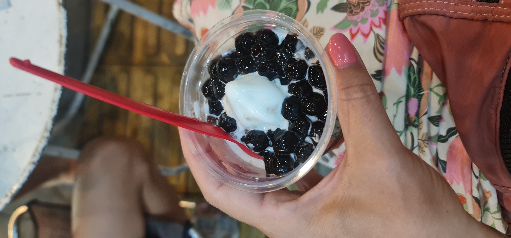 A person's hand holds a clear plastic cup filled with a dessert, likely in a cafe or dessert shop in Đà Nẵng, Vietnam. The dessert consists of a generous portion of white ice cream or creamy base, heavily topped with numerous small, dark, glossy tapioca pearls (boba). A red plastic spoon, faintly branded "Dairy Queen," is nestled in the dessert, indicating the person is either preparing to eat or is in the process of enjoying it. The hand has a visible finger with pink nail polish. In the blurred background, there's a patterned fabric with a floral or leafy design in shades of pink, green, and white, possibly part of clothing or a bag. Adjacent to this, a reddish-brown textured fabric object with visible stitching is present, which could be part of another bag or garment. The overall setting appears casual, with soft, ambient lighting. The specific time of day is not discernible.