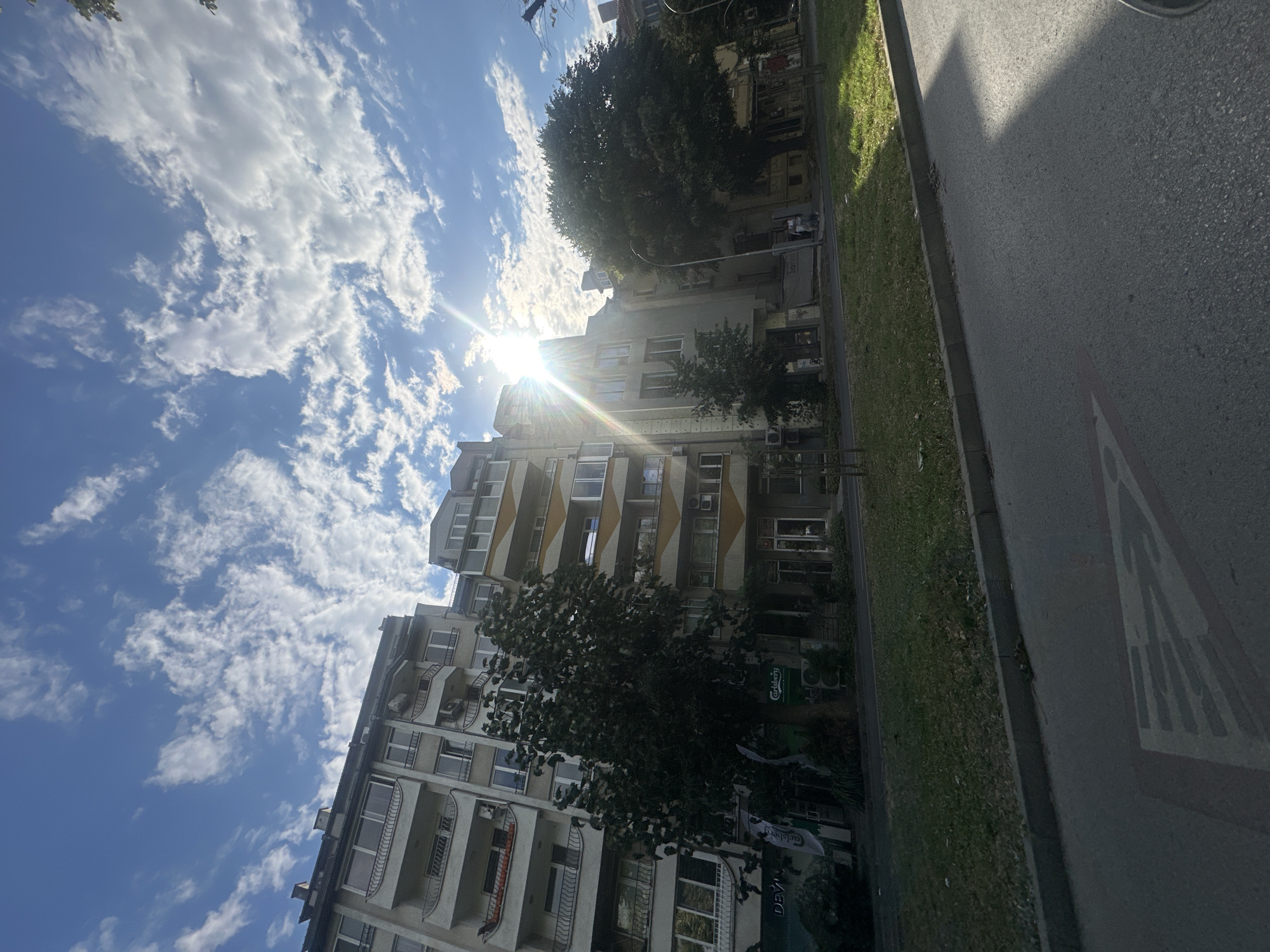 The image captures a sun-drenched street scene with apartment buildings lining the right side. The sky above is a vibrant blue, dotted with fluffy white clouds, and the sun shines brightly, creating lens flares and casting shadows. In the foreground, a grey asphalt road occupies the bottom half of the frame, with a white road marking visible on the left. A strip of green grass and a sidewalk separate the road from the buildings. The apartment buildings themselves are a mix of architectural styles, with balconies, windows, and air conditioning units visible. Some buildings have trees in front of them, and there appears to be some commercial activity on the ground floors. The overall impression is a typical urban environment on a clear, sunny day.