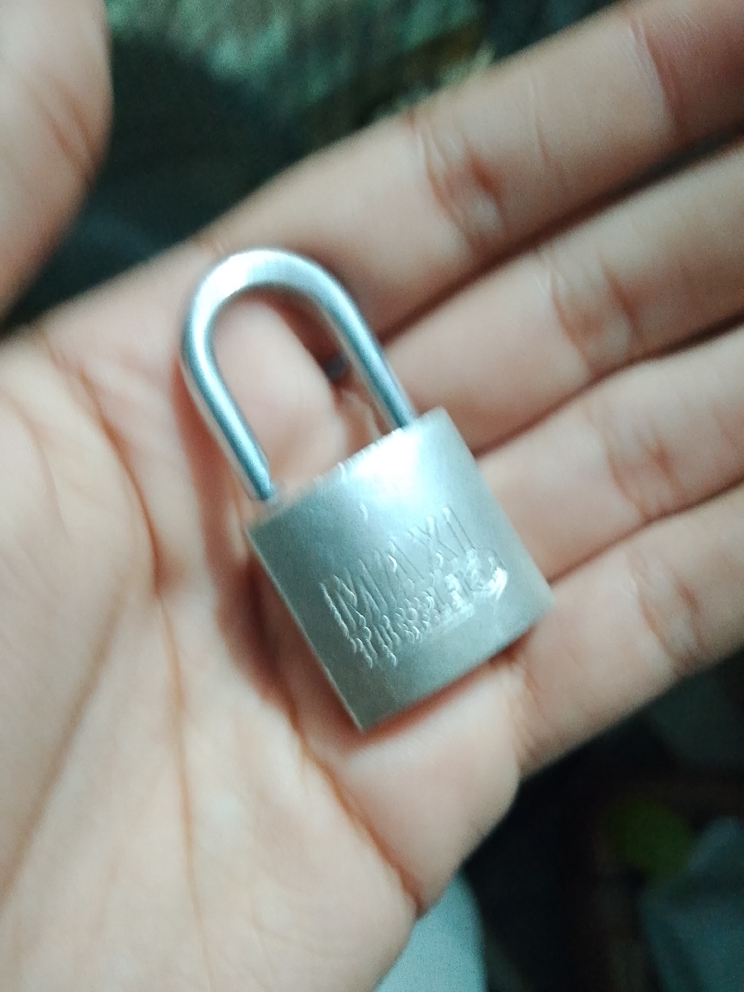 The image is a close-up, out-of-focus photograph of a small, silver-colored padlock being held in a person's palm. The padlock has some embossed lettering on its body, which is partially obscured and difficult to read due to the image quality. The person's hand is open, with the padlock resting in the center. The background is blurred and dark, suggesting an indoor setting with low lighting. There are no identifiable landmarks or context that would indicate a specific location within Santa Ana, Venezuela.