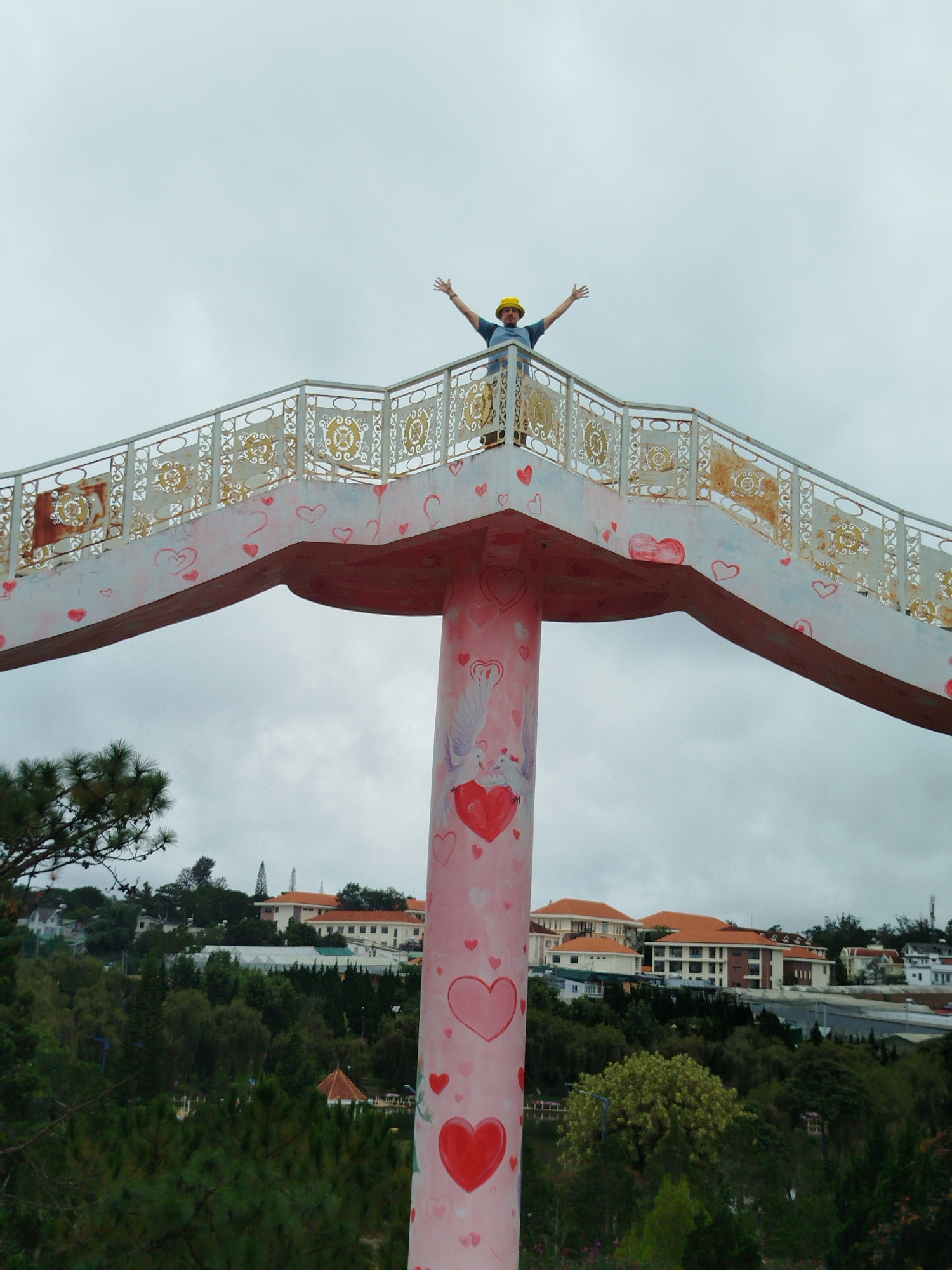 A man wearing a yellow hard hat and a blue shirt stands with his arms outstretched on a pink and white pedestrian bridge decorated with hearts. The bridge has an ornate railing with gold accents. Below the bridge, a cityscape with orange-roofed buildings and lush green trees is visible under a cloudy sky. The bridge's support pillar is also painted pink and adorned with hearts and a depiction of two doves on a heart. The scene appears to be a scenic viewpoint in Lâm Viên - Đà Lạt, Vietnam.