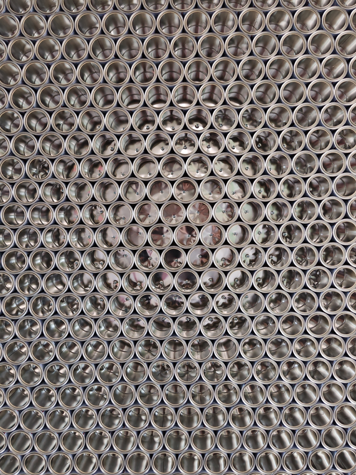 The image displays a large-scale architectural or artistic installation in the city of Gabrovo, Bulgaria, composed entirely of numerous identical or nearly identical silver-colored metal cans. The cans, possibly industrial-grade paint or food containers, are arranged horizontally and tightly packed in a staggered, interlocking grid formation, presenting their open ends directly towards the viewer. Each can features a cylindrical, highly reflective metallic interior, with variations in light and shadow accentuating their depth and the subtle circular patterns or indentations on their inner base. Several cans exhibit small, dark, circular elements resembling fasteners or screws on their internal bottom surfaces. The overall metallic surface gleams under a bright, even illumination. No individuals, specific actions, or interactions are visible within the static scene. The arrangement forms an extensive, textured metallic wall or screen.