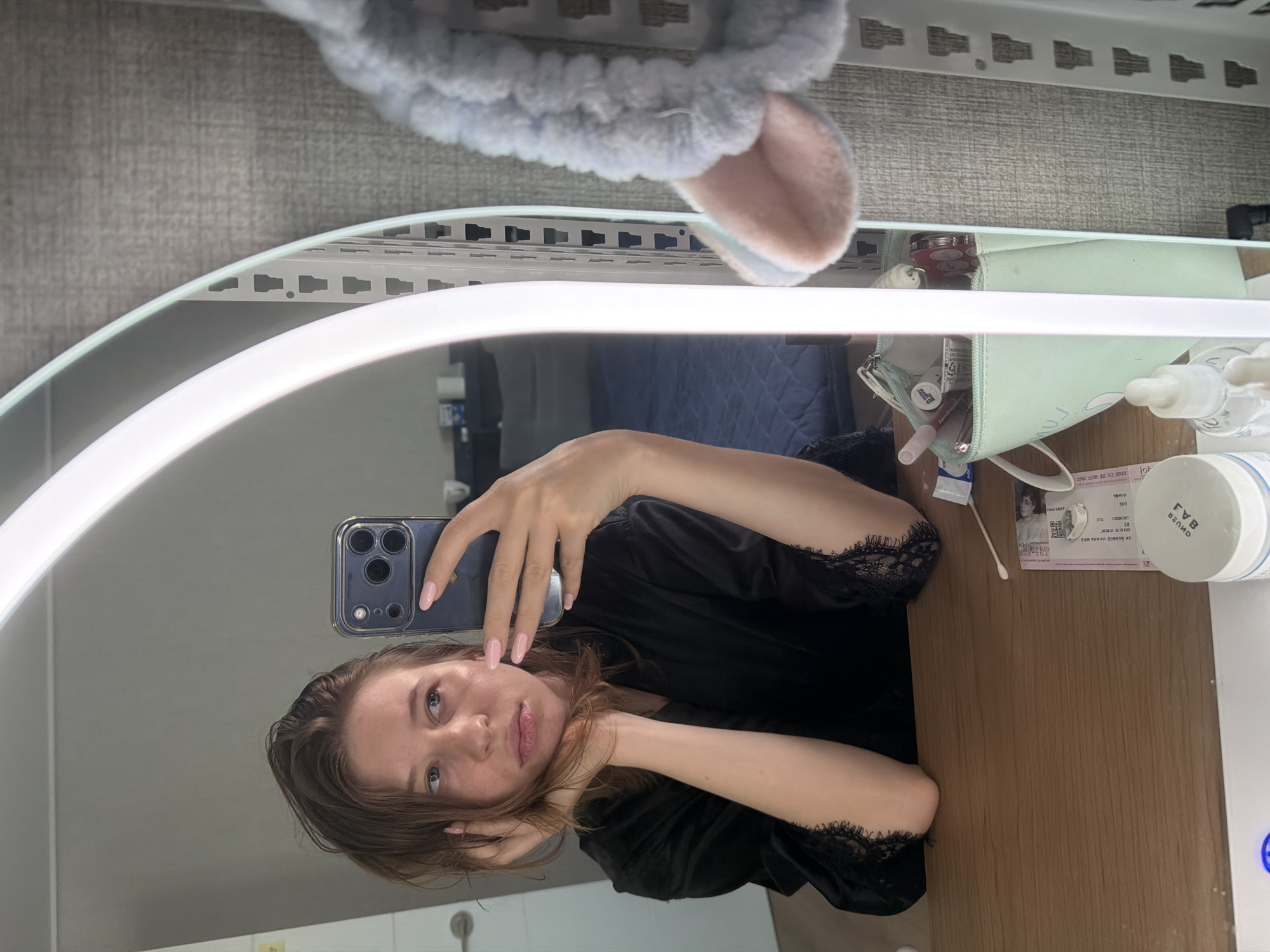 A young woman is taking a mirror selfie in a bedroom. She is wearing a black robe with lace trim and has her hair down. She is holding a smartphone in her right hand, and her left arm is resting on her head. The mirror is illuminated by a bright, curved light. On the table in front of the mirror are several cosmetic items, including a makeup bag, a jar of cream, and a bottle with a dropper. In the background, a bed is visible. The scene is indoors, likely in a domestic setting, and given the context provided, it is located in Dongducheon-si, South Korea.
