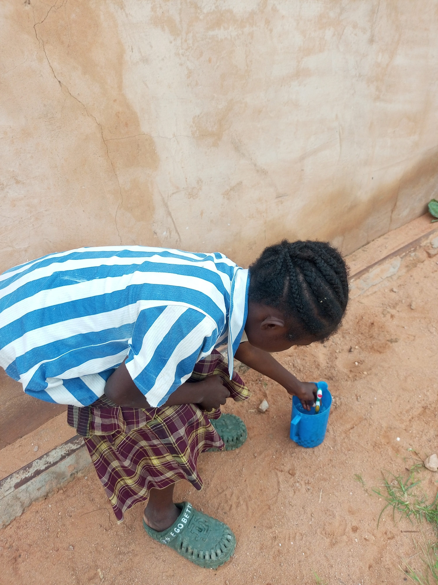 In Tempure, Nigeria, a young child is depicted outdoors, bent over a blue plastic cup. The child is wearing a blue and white striped shirt and a plaid skirt. Green sandals with the inscription "EGOBETTER" are on their feet. The child's right hand is reaching into the cup, where a toothbrush with a white and red handle is visible. The ground is sandy, and a rough, beige wall with cracks forms the background. A concrete ledge runs along the base of the wall.
