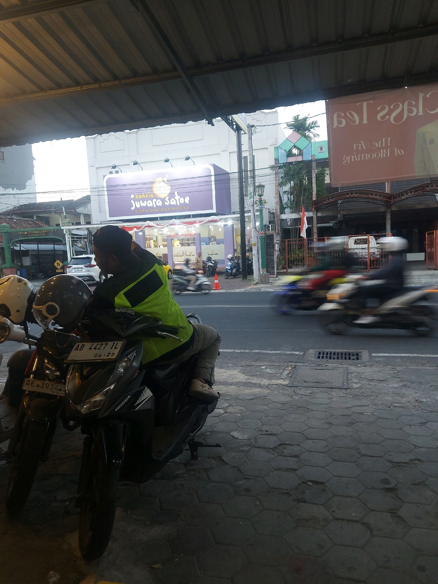 A man wearing a bright green vest and dark pants sits on a black scooter at the side of a street in Banguntapan, Indonesia. The scooter has a license plate that reads "AB 4427 IL 04-23". Two helmets, one white and one gray, are attached to the handlebars of the scooter. In the background, a white building with a purple sign that says "Juwara Satoe" is visible. Across the street, several other scooters are in motion, blurred due to their speed. The street is paved with hexagonal concrete blocks. The scene appears to be in an urban environment, likely during the late afternoon or early evening, given the lighting conditions.