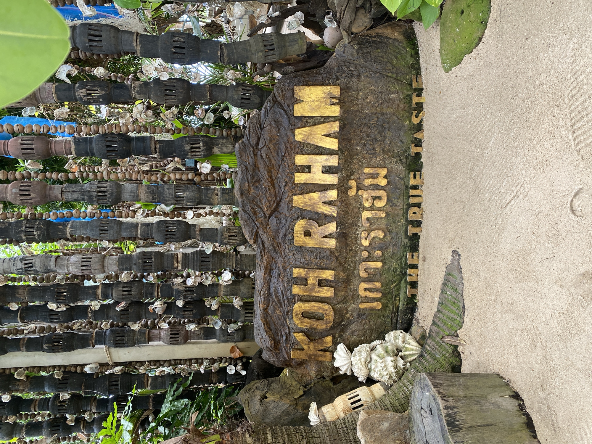 In ตำบลเกาะพะงัน, Thailand, a weathered stone sign is prominently displayed in a sandy outdoor area, likely a resort or restaurant. The sign bears the inscription "KOH RAHAM" in raised golden letters, with Thai characters below it, possibly reading "เกาะราฮัม." At the bottom of the sign, also in golden lettering, are the words "THE TRUE TASTE." The sign itself is textured and appears to be made of a dark, rocky material with moss or algae growing on it.
Behind the sign, a decorative backdrop consists of numerous vertical elements, appearing to be dark wooden posts intricately carved and strung with natural materials such as shells and beads, creating a rustic and bohemian aesthetic. Lush green foliage is visible peeking through the decorative elements, suggesting a tropical environment.
In the foreground, the sandy ground is smooth and undisturbed, with a few scattered decorative items including a large, rough-hewn wooden stump to the left, and a carved decorative element resembling a shell or coral structure. The lighting suggests it is daytime, with natural light illuminating the scene. No people are present in the image. The overall impression is one of a serene, nature-inspired setting designed to welcome visitors.