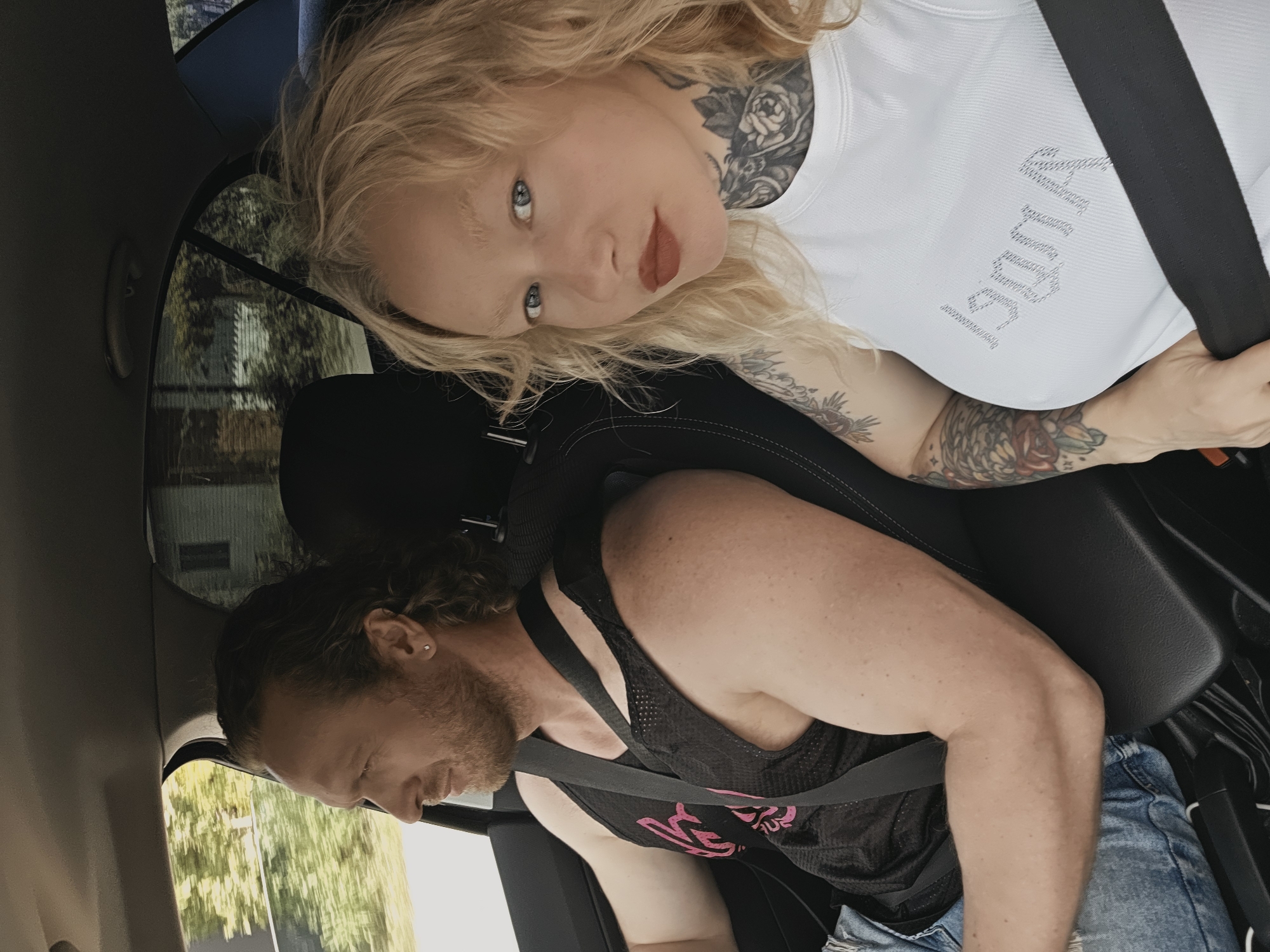 Two individuals are positioned inside a vehicle during daytime.
The driver, an adult male, is seated on the left, oriented towards the driver's side window, suggesting the vehicle is in motion. He has long, wavy, dark hair, a light-colored beard, and a small stud earring in his left ear. He is wearing a black mesh tank top with a pink design partially visible on the chest, and light blue ripped jeans. A black seatbelt is diagonally across his torso. His right arm is visible, showing muscular definition.
The passenger, an adult female, is seated on the right, facing the camera. She has long, wavy, blonde hair, light-colored eyebrows, blue eyes, and red lipstick. She is wearing a white ribbed tank top with silver-toned, possibly rhinestone, lettering on the chest (partially legible as "UMA" or "PUMA" in reverse). Visible tattoos adorn her neck (a floral and skull motif) and her left arm (including a bird-like figure and a rose). A black seatbelt is diagonally across her torso and horizontally across her lap. Her right hand is positioned near the seatbelt buckle.
The car's interior features dark upholstery for the seats and ceiling. A dark center console is visible between the two occupants. Through the windows, blurred green foliage and the structure of a light-colored building are discernible, indicating the vehicle is moving through an outdoor environment, likely a residential area or suburban road.