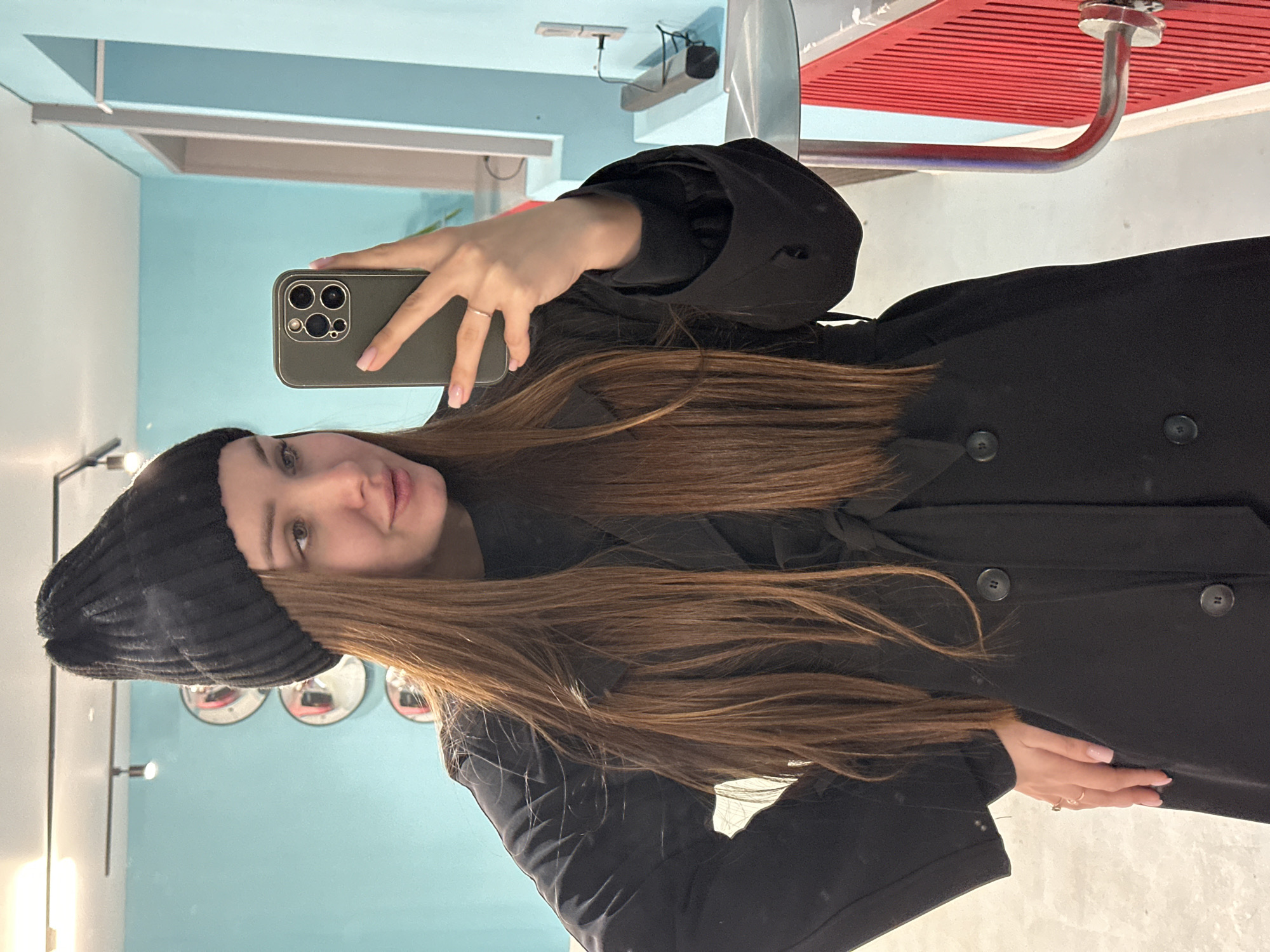 An indoor mirror selfie captures a young woman from the chest up, centered in the frame, in what appears to be Chaikovsky, Russia. She holds a smartphone with a dark green case (resembling an iPhone with prominent camera lenses) to take the photo, partially obscuring her face. Her long, straight, medium brown hair cascades over her shoulders and down her front. She is wearing a black ribbed beanie and a black long-sleeved coat or jacket with visible buttons. On her right hand, which holds the phone, a simple silver-colored ring is visible, and on her left hand, which rests near her waist, a gold-colored ring is present. Her nails have a light, natural polish.
The woman looks towards the camera with a subtle smile and a calm expression. The background, visible in the mirror's reflection, primarily features a light blue wall. In the upper right of the reflection, a red slatted structure, possibly a ceiling vent or decorative panel, is visible, along with metallic piping or fixtures. The upper left shows another shelf-like structure against the blue wall. In the lower left, three circular mirrors are mounted on the blue wall, reflecting parts of the room and creating a decorative effect. The lighting is bright and even, suggesting an indoor setting like a café, changing room, or a well-lit public space. There is no visible text in the image.