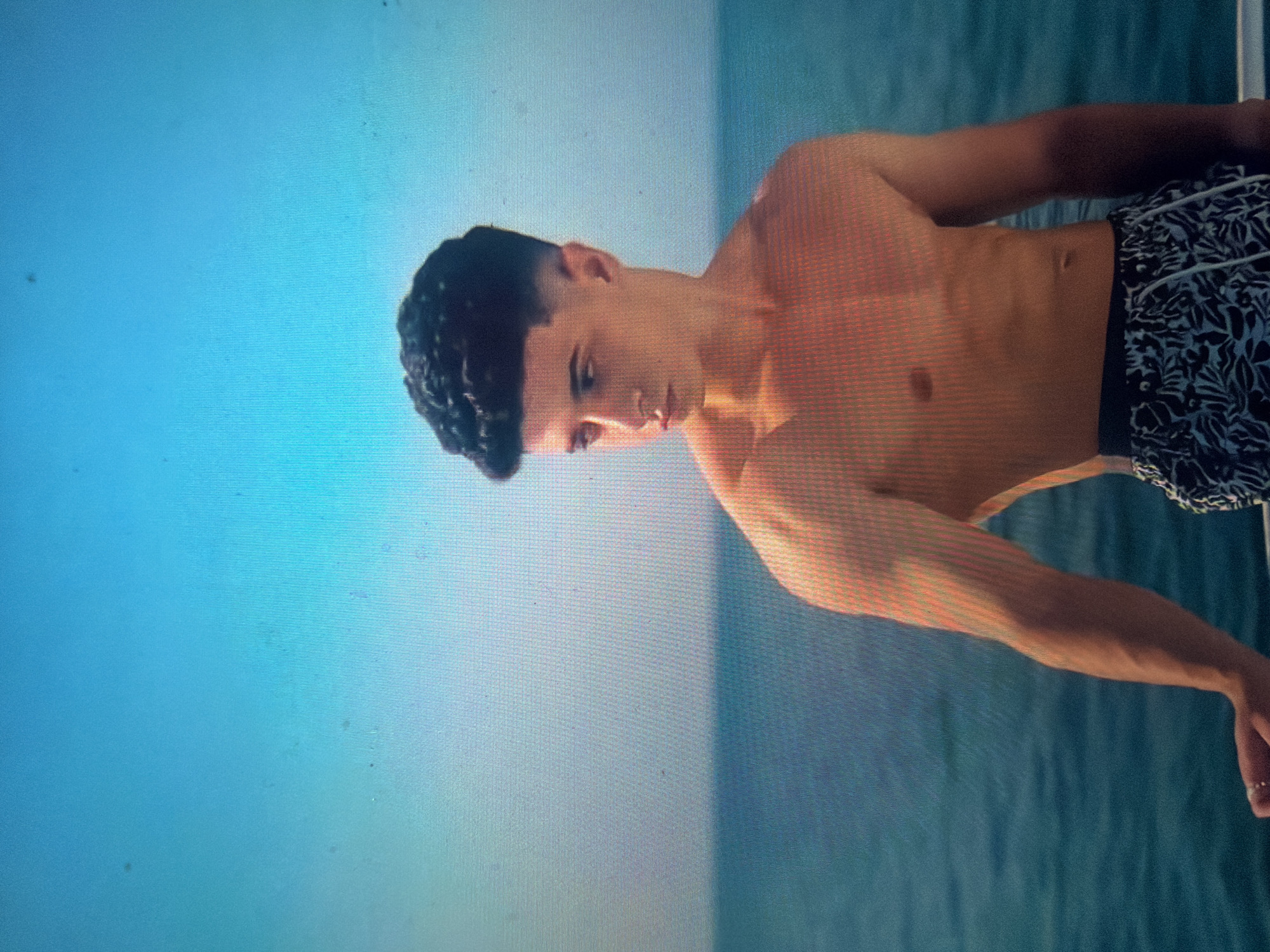 The image displays a young adult male, shirtless, standing on what appears to be a boat or waterside structure. He has dark, curly hair with closely shaven sides and a more voluminous top. He is facing left from the viewer's perspective, looking slightly downwards and away, with his head turned approximately 45 degrees from the camera. His left hand, adorned with a metallic ring on the ring finger, rests on a thin, dark railing or edge in the bottom-left foreground. He is wearing dark swim trunks with a white or light-colored leaf or floral pattern and a white drawstring. The background consists of a vast expanse of calm, blue-green water meeting a clear, light blue sky at the horizon, indicating an open-air environment. A prominent grid pattern of pixels is visible across the entire image, suggesting it is a photograph captured from a digital screen rather than a direct photograph of the scene itself. No specific landmarks or details within the image indicate the city of Shumen, Bulgaria; the environment is solely open water and sky.