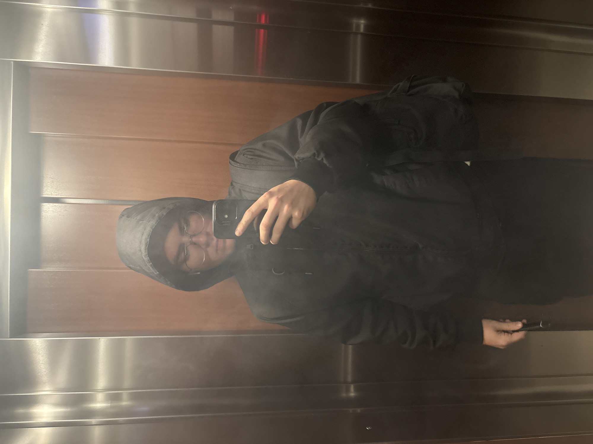 The image captures a mirror selfie of a person inside an elevator. The subject, an individual wearing a dark, possibly hooded jacket and eyeglasses, is visible from roughly the chest upwards, holding a dark-colored smartphone with their right hand to take the photo. A dark backpack is faintly discernible on their back, blending with their jacket.
The elevator's interior walls are a mix of materials. The central section features horizontal panels of light brown wood, exhibiting a distinct grain pattern. Above and below these wooden panels are highly polished metallic surfaces, likely stainless steel, which are very reflective, mirroring the person and internal lighting. The overall lighting appears to be internal to the elevator, creating clear reflections across the various surfaces.
The primary activity is the taking of a mirror selfie. No specific emotions are evident from the person's expression, which is focused on the task. There is no visible text within the image. The location context provided for the image is Plovdiv, Bulgaria.