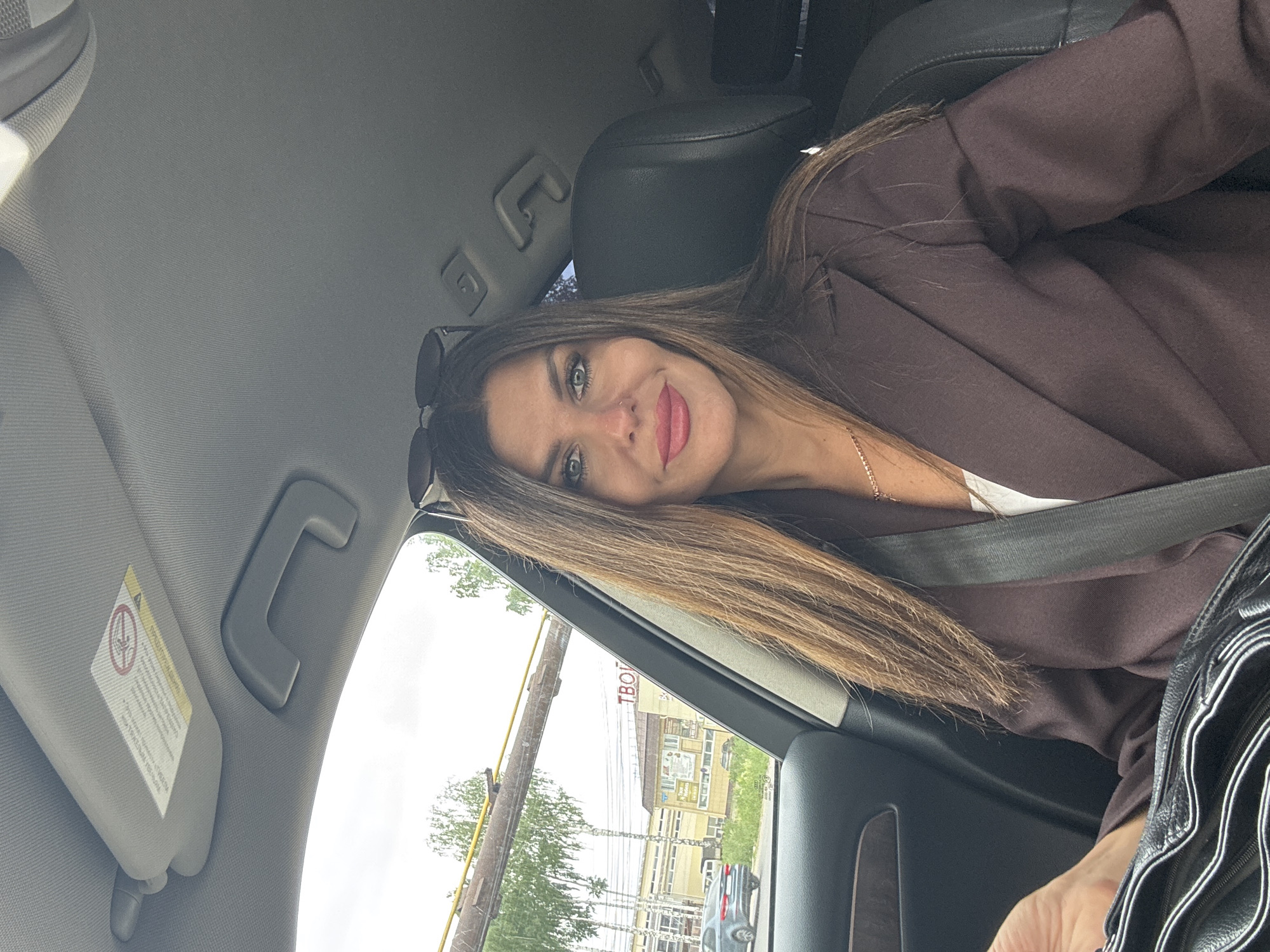 A woman with long, brown hair and makeup, wearing a brown blazer and a white top, is seated in the driver's seat of a car. She is looking at the camera, holding a black purse. Outside the car window, trees, buildings, and a road are visible. One building has signage with Cyrillic letters that partially reads "ТВОЙ". The image appears to be taken in Chaikovsky, Russia.