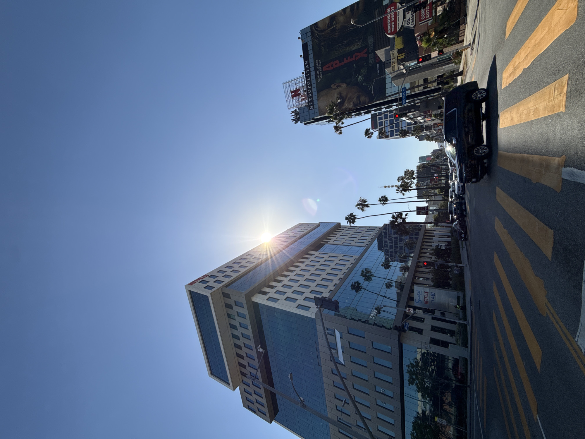 This image is a ground-level view of a city street in Los Angeles, USA, looking upward at a modern building with a mix of glass and concrete facade. The building's name, "NETFLIX KTLA 5," is visible on its lower section. The sun is shining directly above the building, creating a starburst effect. Palm trees line the street, adding to the Los Angeles cityscape. On the right of the building, cars are parked in front of the cross walk on the right. The Billboard displays "APEX" movie with Charlize Theron and Taron Egerton. Additional billboards and signs are visible, including "AMERICAN TIRE DEPOT." The sky is clear and bright blue, suggesting a sunny day.