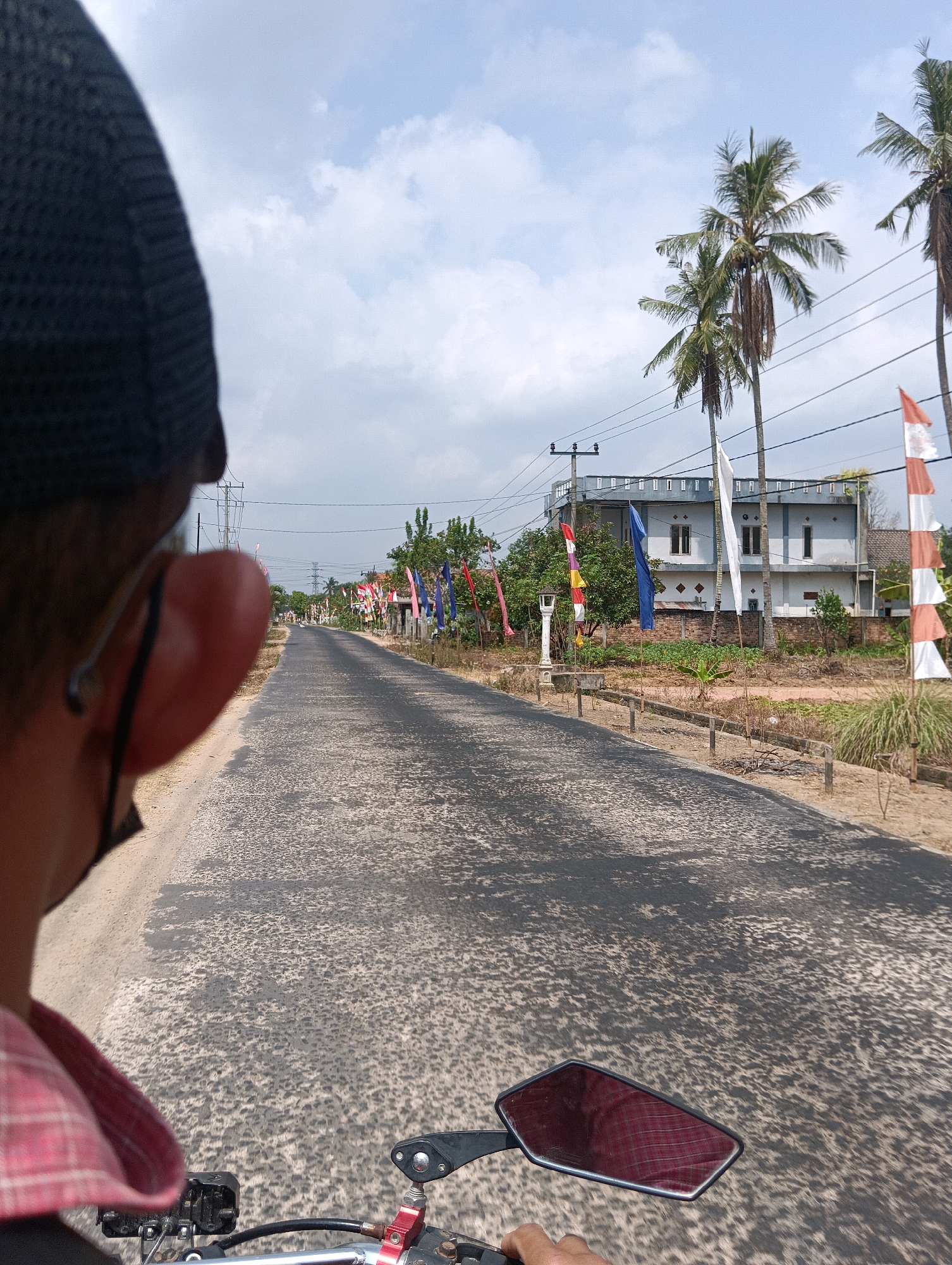 This is a first-person perspective, outdoor, daytime, eye-level shot of a person riding a motorcycle on a paved road. The rider is wearing a dark cap and a red and white checkered shirt. The road is lined with trees and flags, and there are buildings in the background. The sky is blue with some clouds. The location is the city of Nusa Bali, Indonesia.