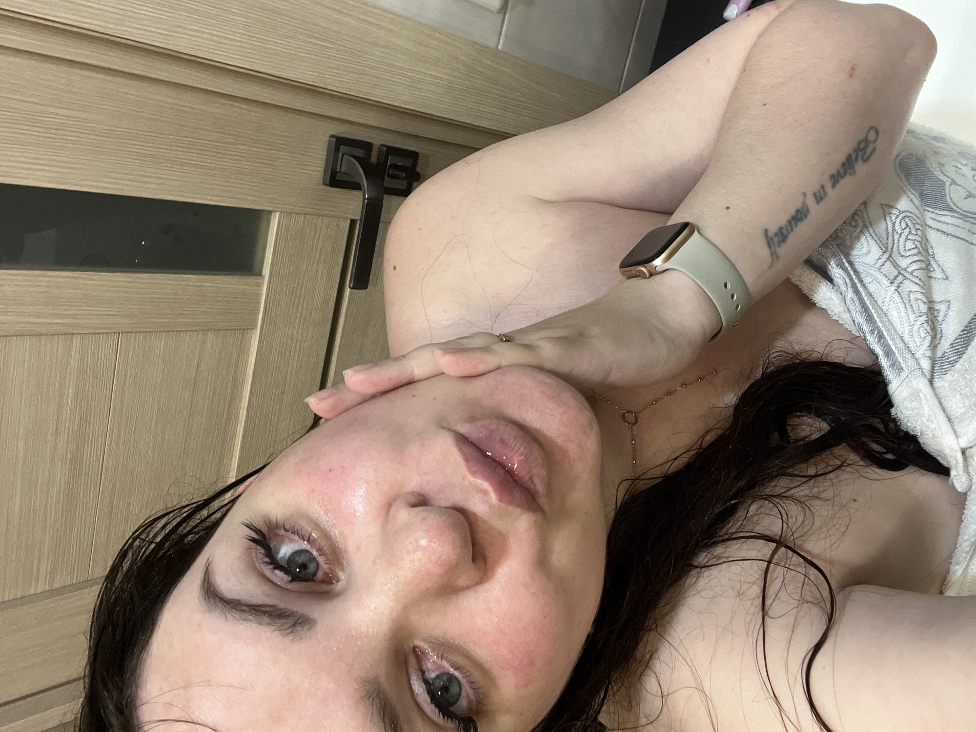 A close-up, selfie-style image shows a woman with dark hair and fair skin. She is facing the camera, with her right arm raised, hand touching her face. A towel covers part of her right shoulder. A smartwatch is visible on her right wrist, and a tattoo is on her right forearm. A light-colored wood paneled door with a dark handle is in the background. The location is not determined.