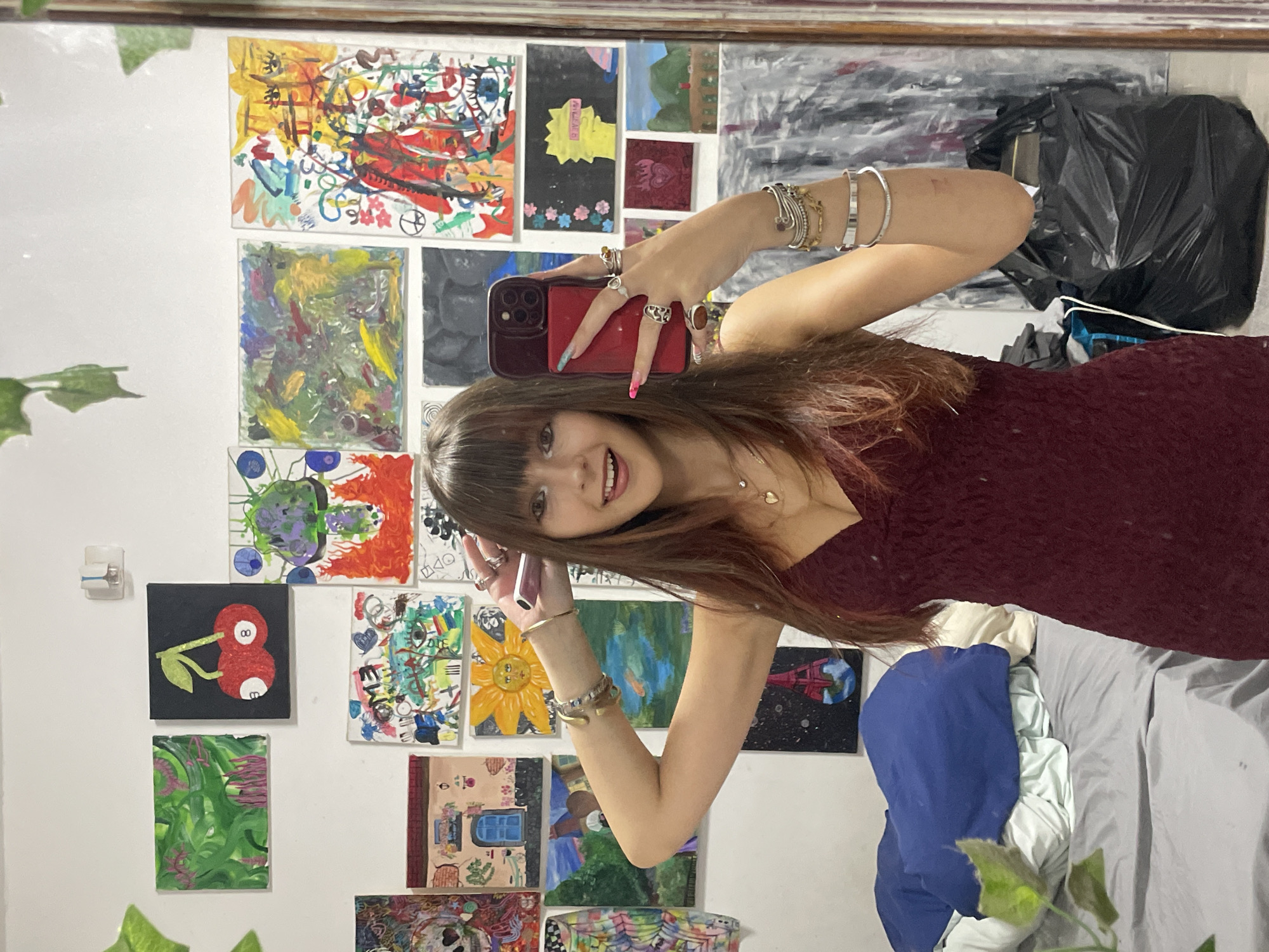A young woman with brown hair and bangs, wearing a dark red lace dress, poses for a selfie in a bedroom. She has a phone in her right hand, and her left arm is bent upwards, showcasing several bracelets on her wrist. The wall behind her is covered with numerous colorful paintings. To the left, a bed is partially visible with rumpled blue and white bedding. In the lower right corner, a large black trash bag stands near a reflective surface, likely a mirror. The scene is likely within Raanana, Israel, given the context.