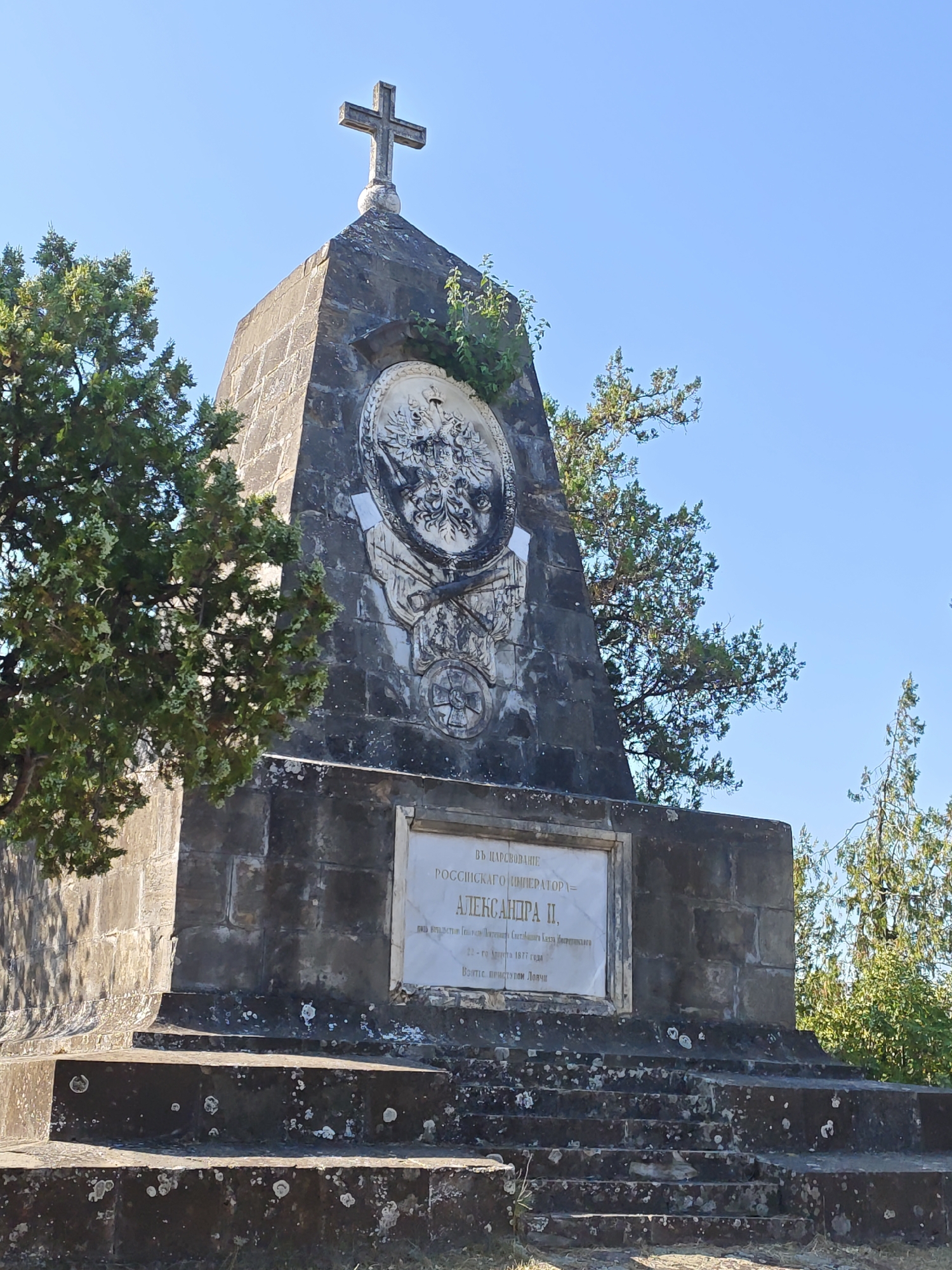 The image shows a stone monument topped with a cross, set against a clear blue sky. The monument is weathered and appears to be made of rough-hewn stone blocks. A circular relief carving, featuring a double-headed eagle, is prominently displayed on the front of the monument, partially obscured by greenery. Below this, a white plaque with inscribed text is affixed to the monument. The monument stands on a tiered base, also made of stone, with visible signs of age and wear. Several trees with green foliage surround the monument. The location is identified as the Monument to the Russian Emperor Alexander II in Lovech, Bulgaria.