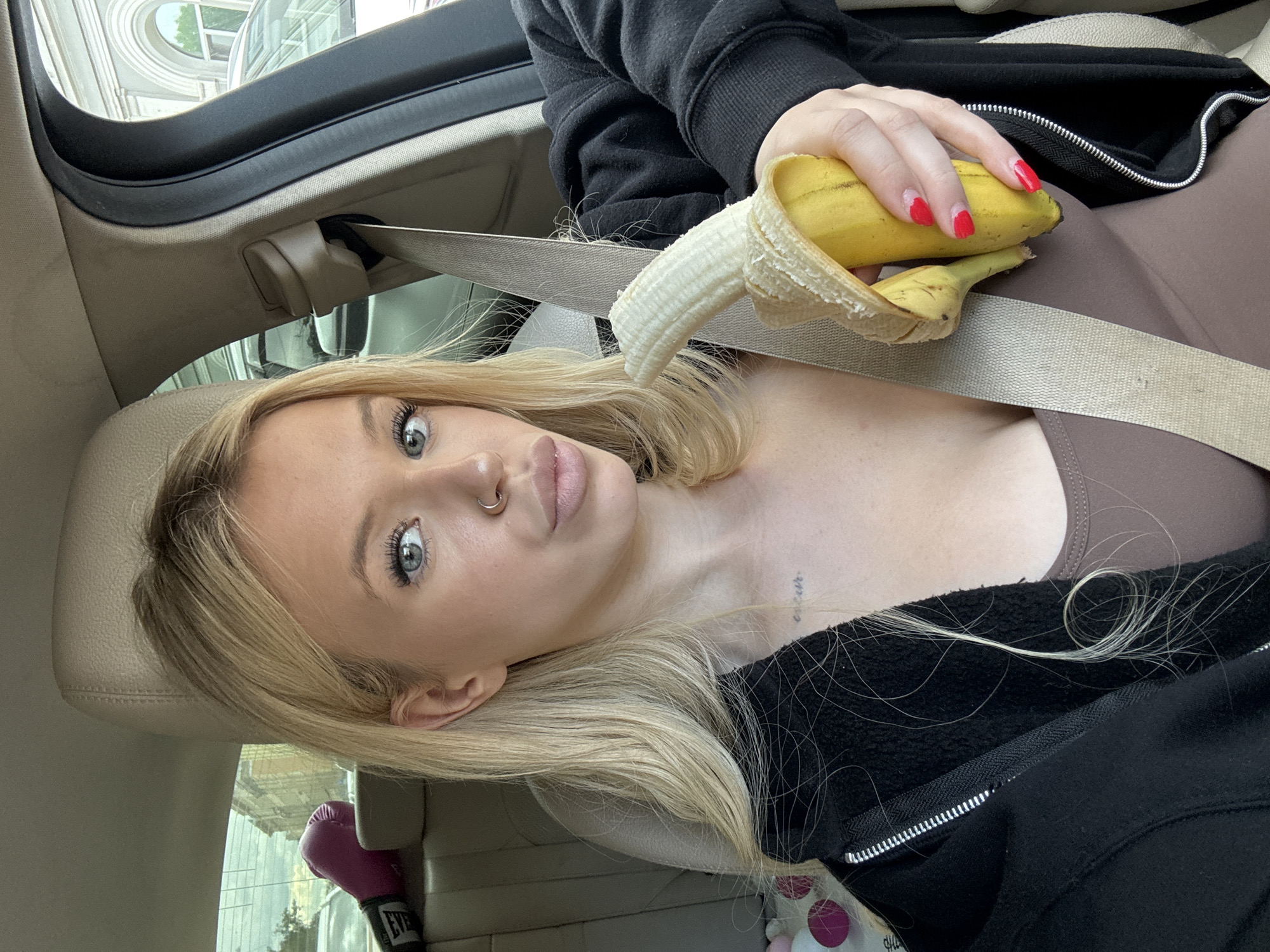 This media file captures a close-up, Dutch angle shot of a young woman with blonde hair and light blue eyes sitting in the passenger seat of a car. She is wearing a brown tank top and a black zip-up hoodie. Her right hand is holding a partially peeled banana, with her fingernails painted red. A silver nose ring is visible on her nose. A delicate tattoo can be seen on her collarbone. The car interior is visible, including a beige seatbelt, a beige headrest, and part of the dashboard. Through the car window, a building with classical architecture and an arched window is visible, suggesting an urban environment. In the background, a pair of pink boxing gloves with "EVERLAST" printed on them can be seen. The lighting suggests it is daytime. The woman's expression is somewhat direct, with a slight tilt of her head. The overall impression is a casual, personal moment captured in a car. The text "EVERLAST" is visible on the boxing gloves.