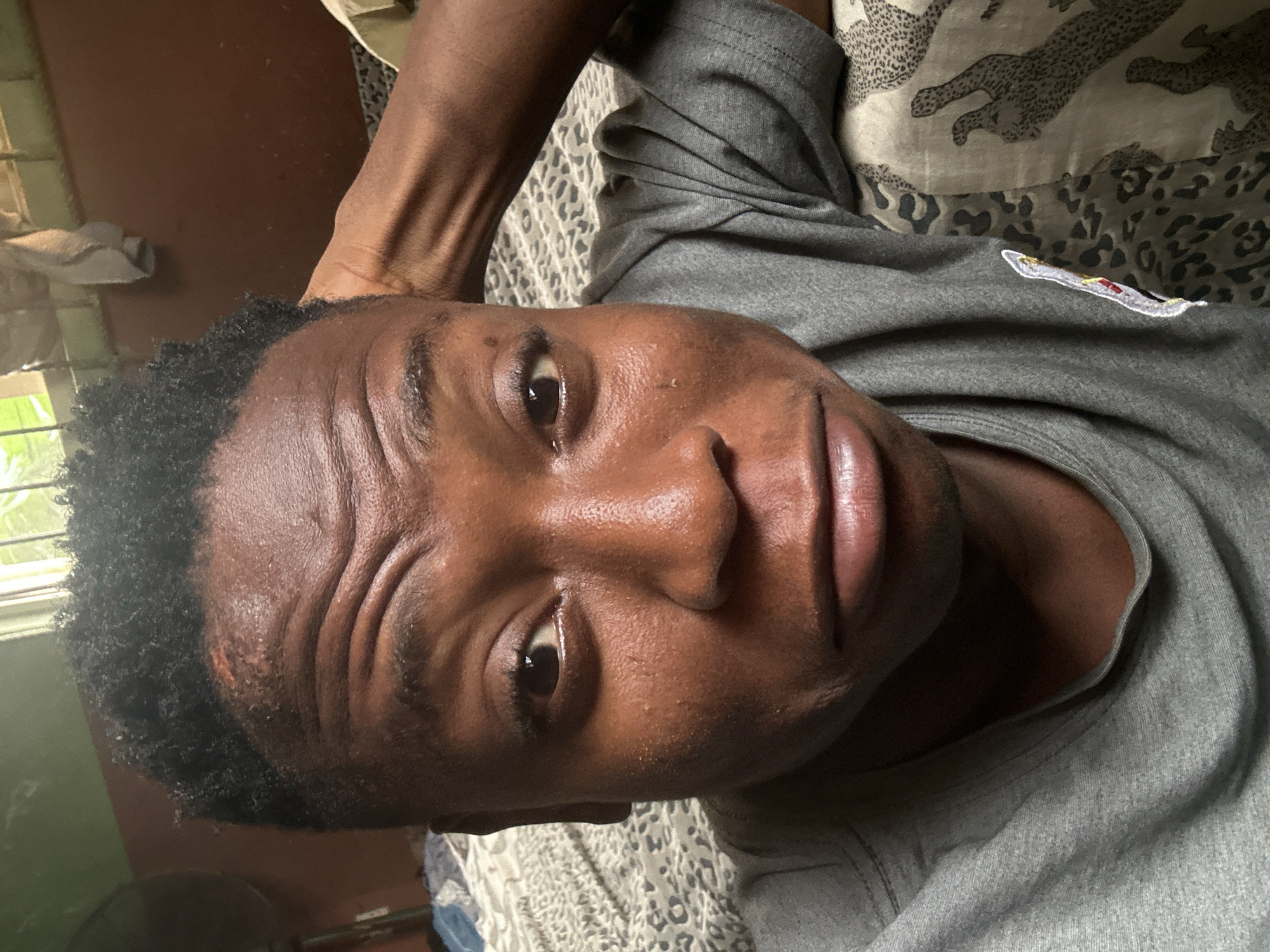 A close-up selfie shows a Black individual resting their head on their hand, with their arm bent and behind their head. The individual appears to be looking at the camera with a slightly pensive expression, with furrowed brows and a downturned mouth. They are wearing a gray t-shirt. The background is dimly lit, with a patterned bedspread and a glimpse of a window with bars. The scene is likely indoors, possibly in a bedroom. The presence of a fan is visible in the far left of the image. The overall impression is one of casual repose. The individual's location in Ikorodu, Nigeria, is confirmed through visual cues present in the background, such as the style of the window and the general domestic setting, which are consistent with residential areas in that region.