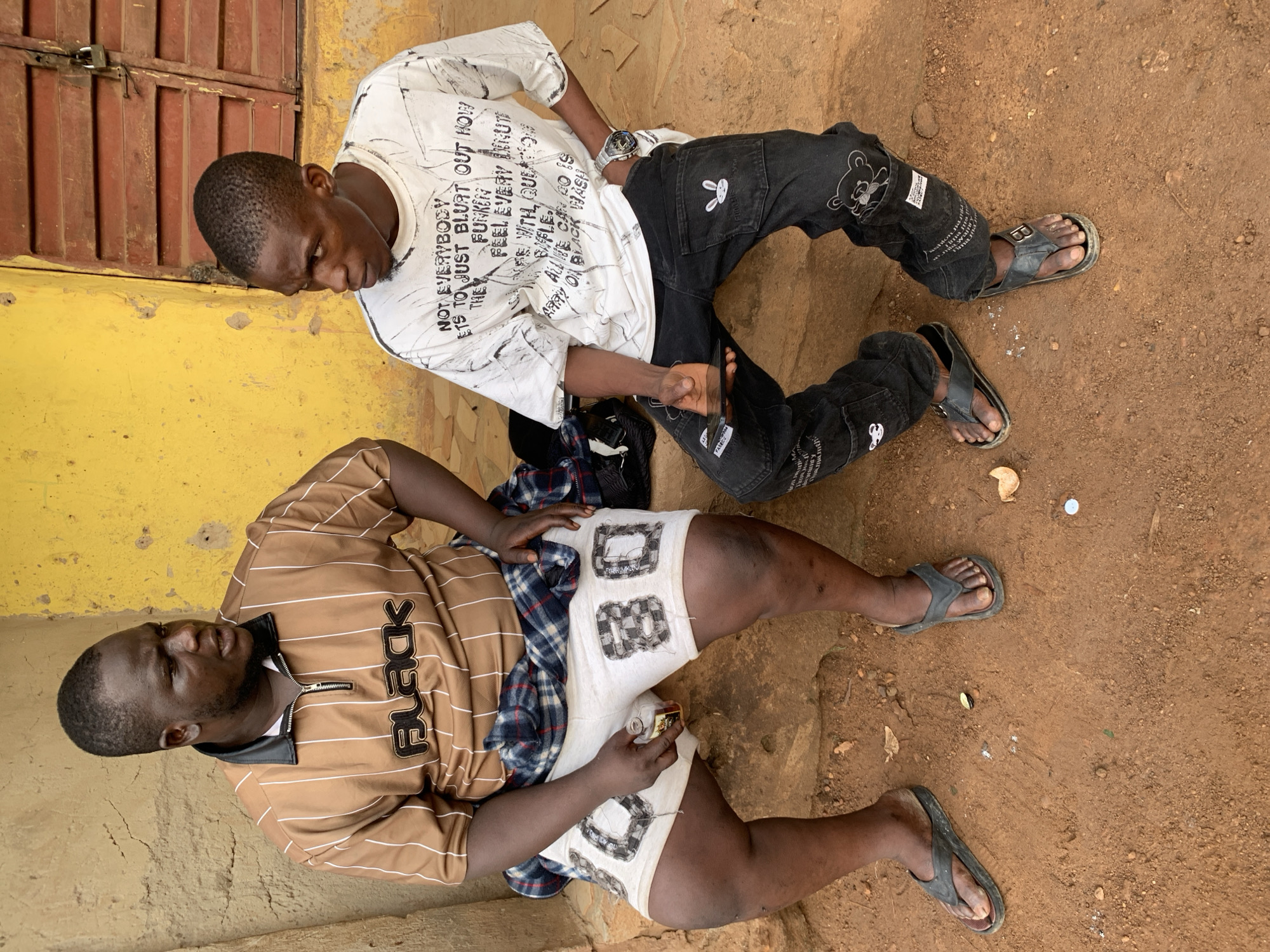 Two adult males are seated on a reddish-brown dirt ground in Akwanga, Nigeria, against a backdrop of a yellow-painted wall and a brown corrugated metal structure. The image appears to be taken during the daytime under natural light.
The man on the left wears a brown and white striped polo shirt with a black collar and a zipper, a white undershirt, and white shorts adorned with block-style printed letters, including a clear "B". A blue and white plaid shirt is draped over his lap, and he holds a small, rectangular metallic object. He wears black flip-flops. Vertical text on his polo shirt reads "FUCK".
The man on the right wears a white long-sleeved t-shirt featuring black abstract patterns and extensive text. The text includes phrases like "NOT EVERYBODY GETS TO JUST BLURT OUT HOW THE FUNKY FEEL EVERY MINUTE ALL I NE CAN DO IS CARRY BLACK HASH". He also wears black jeans with cartoon character patches (a white bunny and a bear) and text, including "FASECO". A silver watch is visible on his left wrist, and he appears to be holding a pair of sunglasses. He also wears black flip-flops.
Both men are seated casually on the ground, possibly resting or engaging in a conversation. The ground is dry, showing small stones and debris.