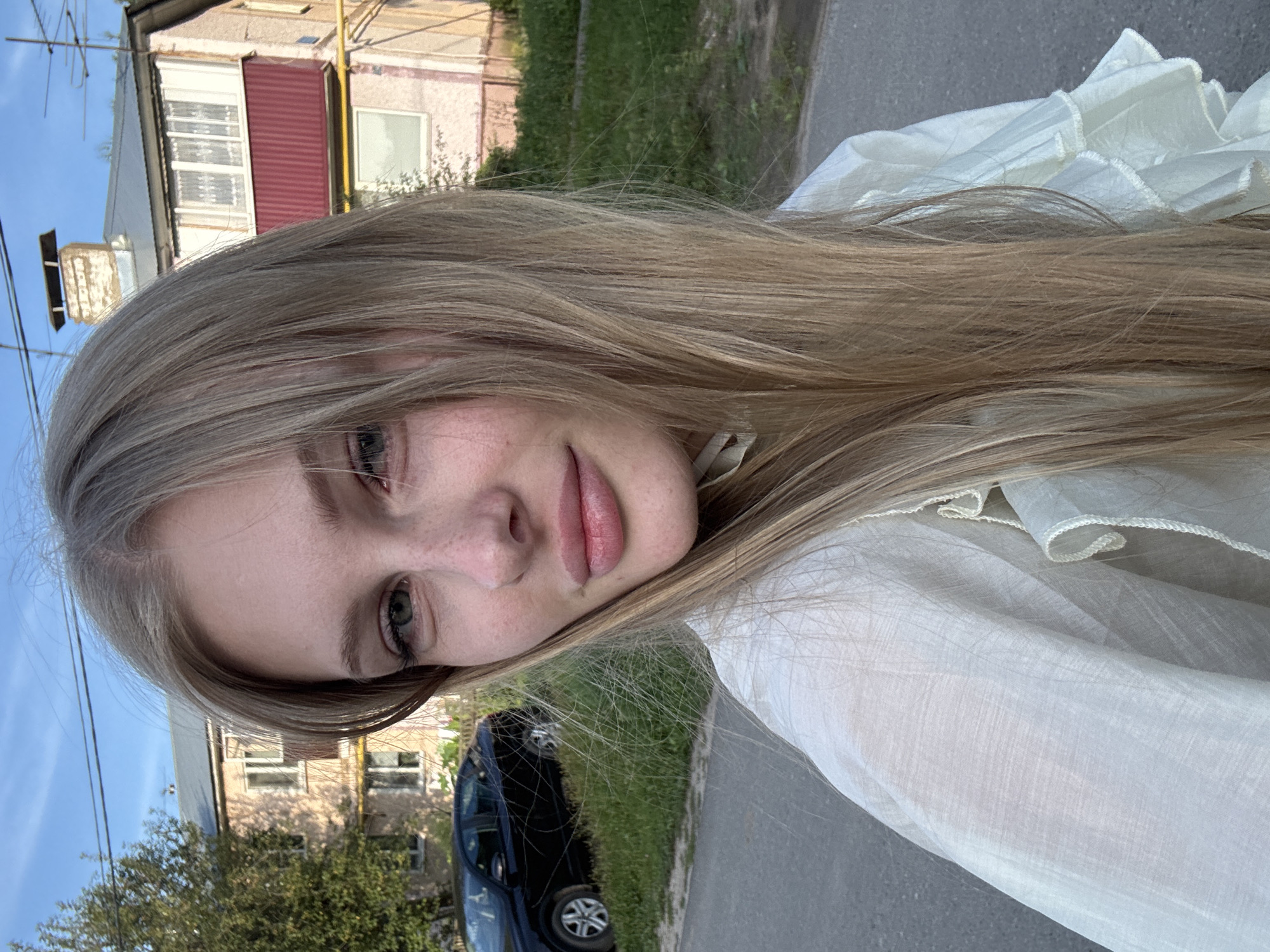 A young woman with long blonde hair and light skin, wearing a white ruffled blouse, stands outdoors. She has freckles on her cheeks and light green eyes, and her lips are lightly colored. Behind her, a black car is parked on the side of a paved road, with a grassy area and a pink multi-story building in the background. The sky is clear and blue with a few wispy clouds. The scene appears to be a residential area, possibly in a city like Kazan, Russia, given the architectural style of the building and the presence of utility poles and overhead wires, common in many Russian cities. The lighting suggests it is late afternoon or early evening.