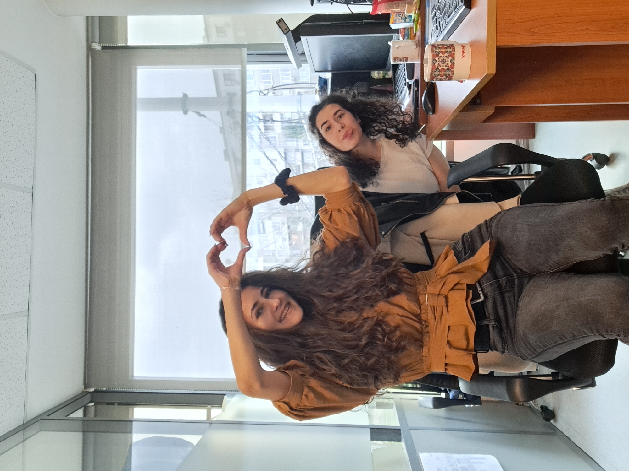 Two women are in an office setting. One woman is in the foreground, making a heart shape with her hands while smiling. She is wearing a brown blouse and jeans. The other woman is in the background, sitting at a desk with a computer and a cup. She is sticking her tongue out playfully. The office has a large window with a view of buildings outside. The exact location in the city of Sofia, Bulgaria, cannot be ascertained from the image.