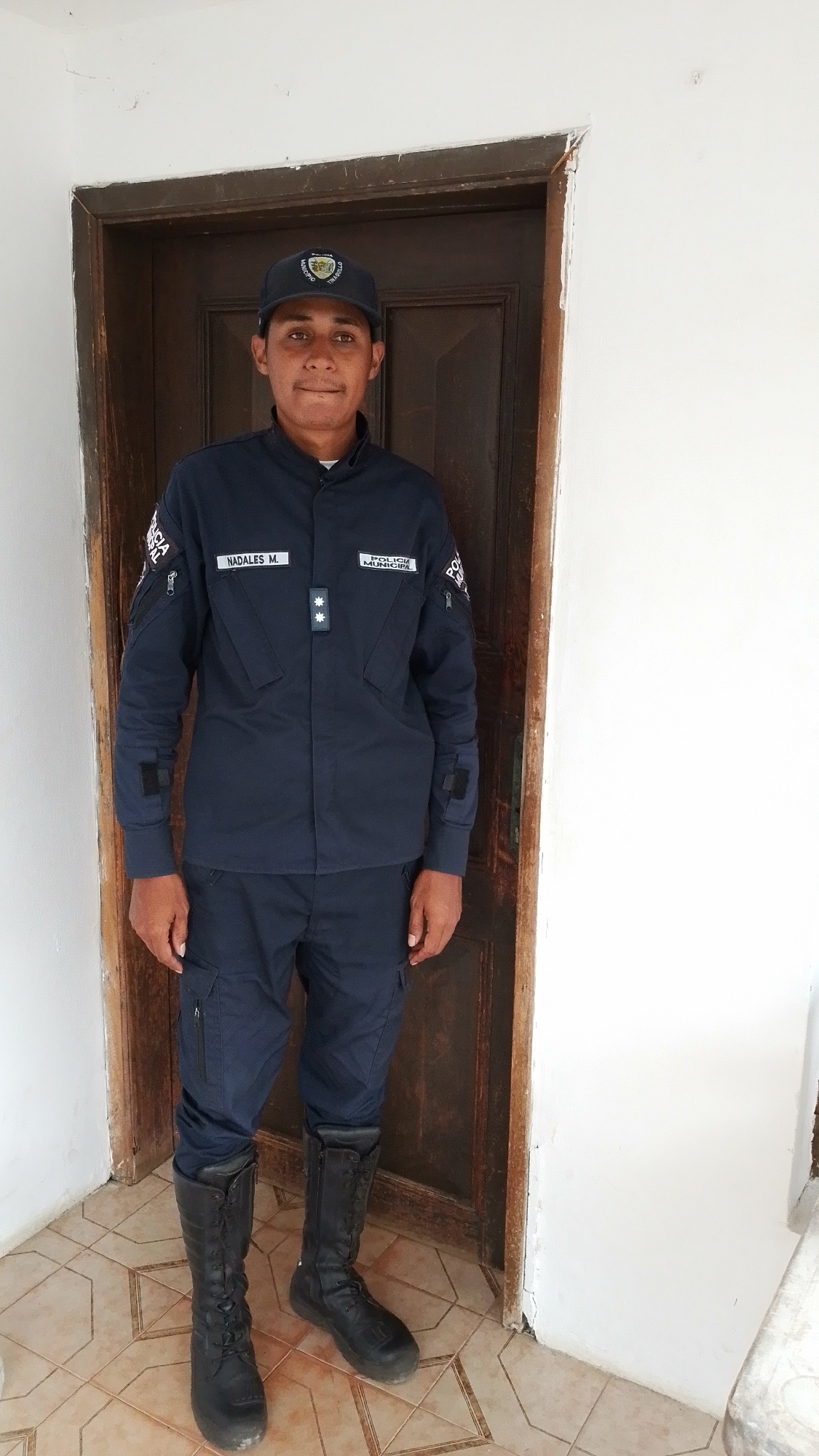 A young man in uniform stands facing the camera, posing in a doorway. He has dark hair, dark eyes, and light brown skin, with a neutral to slightly smiling expression. He wears a dark blue uniform consisting of a long-sleeved shirt and tactical trousers, paired with tall black lace-up boots. A dark blue baseball cap with a circular emblem featuring the word "MUNICIPIO" is on his head.
On the left chest of his uniform, a white name tag reads "NADALES M." On the right chest, another white patch states "POLICIA MUNICIPAL." Below these patches, on a dark blue tab, are two silver stars indicating rank insignia. Patches are also visible on both sleeves, including text that appears to be "POLICIA NACIONAL" or similar.
The man stands centered in front of a dark brown wooden door with a simple frame. To his left and right are plain white walls. The floor is covered with light brown and beige patterned tiles. The lighting suggests it is daytime, casting no harsh shadows. The overall setting appears to be an indoor area, possibly an entrance or hallway in Tinaquillo, Venezuela.