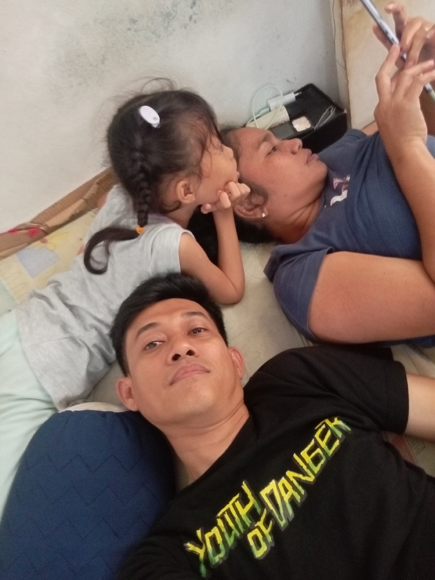 A family, likely residing in Batam, Indonesia, is captured in an informal indoor setting, possibly a bedroom, as suggested by the presence of bedding and a wall that appears to be a partition. A man in the foreground, wearing a black t-shirt with "YOUTH OF DANGER" emblazoned on it, lies on a blue pillow. To his left, a young girl with braided hair and a white hair clip is propped up, her arm resting on her face, indicating a moment of contemplation or rest. Beside the girl, a woman is lying down, engrossed in her smartphone. The background reveals a simple wall and what appears to be a charging device plugged into an outlet. The overall atmosphere suggests a relaxed, domestic scene.