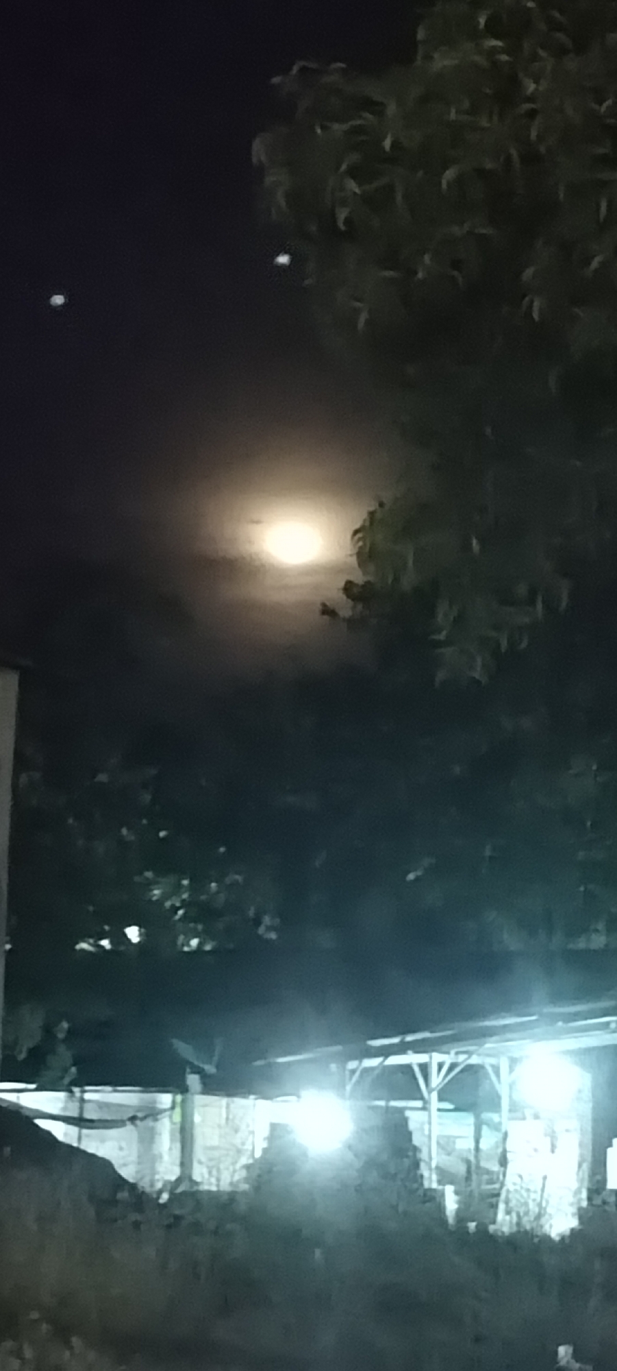 The image shows a night scene in Lhokseumawe, Indonesia. The sky is dark with a bright full moon partially obscured by clouds and foliage. There are two other small illuminated objects in the sky, possibly stars or lights. A tree with dense leaves is visible in the foreground on the right side of the image. Below, there's an illuminated building with a simple frame structure, emitting a bright glow. In front of the building, there is a pile of objects or construction materials on the ground, blending into the darkness. The overall scene is dimly lit by the moon and artificial lights, creating a stark contrast between light and shadow.
