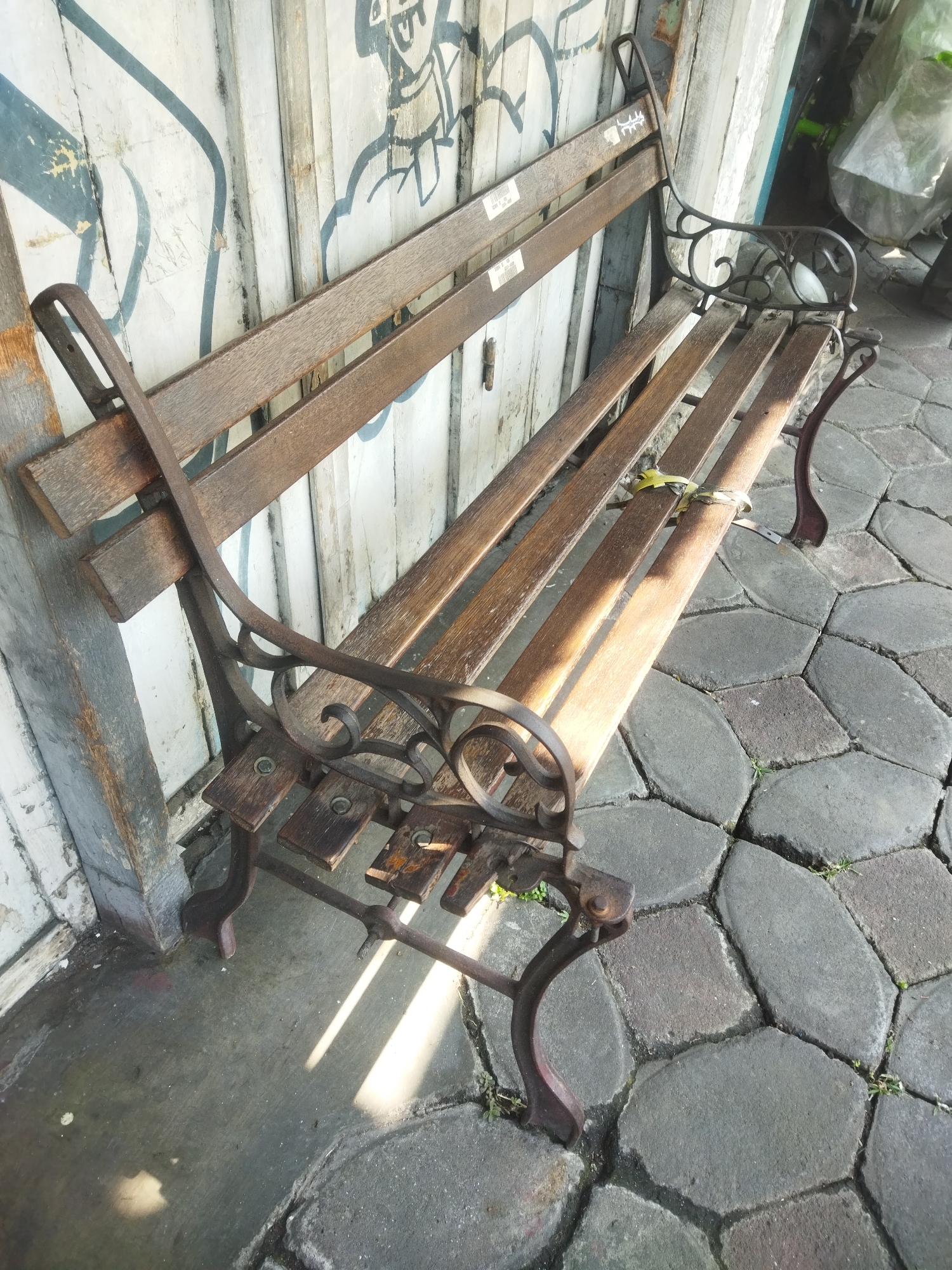 A weathered wooden and metal park bench is situated on a stone-tiled sidewalk. The bench has a metal frame with decorative scrollwork on the armrests and legs. The wooden slats of the seat and backrest appear aged and worn, with some showing signs of repair or modification, such as yellow zip ties holding two slats together. The bench is positioned next to a vertical wooden wall or door, which is covered in graffiti. To the right of the bench, there is a paved area with irregularly shaped gray stones. The ground in front of the bench is a concrete surface, part of which is wet and reflects sunlight. In the background, behind the bench and to the right, there are some indistinct objects covered in plastic sheeting, and a hint of green foliage. The scene suggests an outdoor urban environment. This image was taken in Giripurwo, Indonesia.