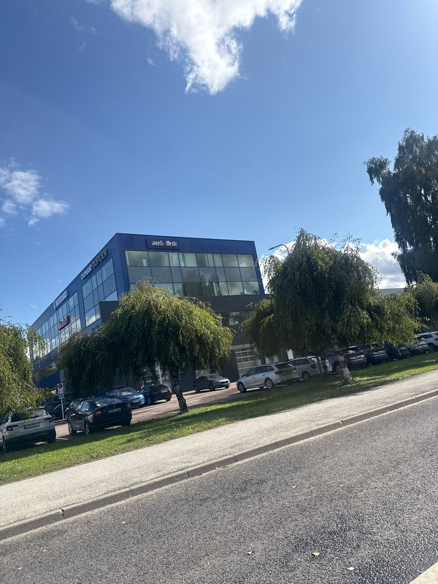 The image depicts a modern, blue office building with large glass windows, situated in a commercial area. The building has signage that reads "OUMAN OU FLEX" and "autolink". Several cars are parked in a lot in front of the building, which is lined with trees with drooping branches. The scene appears to be a daytime shot under a clear blue sky with scattered white clouds. This location is consistent with the office park and business areas found in Tallinn, Estonia.