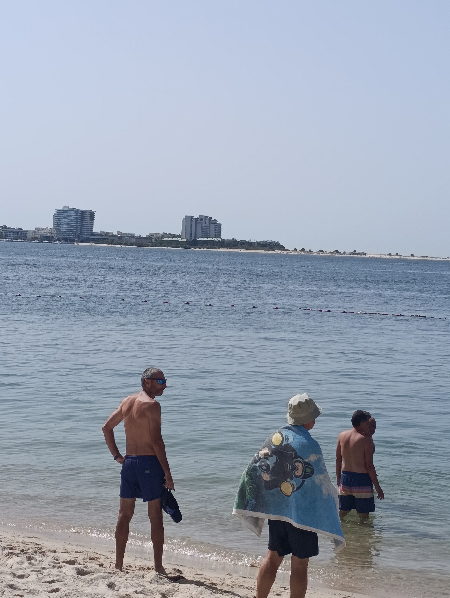 Three men stand in the shallow waters of a beach. The man in the foreground, facing away from the camera and to the right, wears dark blue swim trunks and sunglasses. He holds a black flip-flop in his right hand. To his left, a man with a towel draped over his shoulders, featuring a graphic of a diver, also faces away from the camera. He wears a light-colored hat and dark swim trunks. Further to the right and slightly deeper in the water, a third man faces partially away from the camera and to the left, wearing multi-colored striped swim trunks. The background features the ocean, a sandy beach, and a distant cityscape with modern buildings. The sky is clear and pale blue.