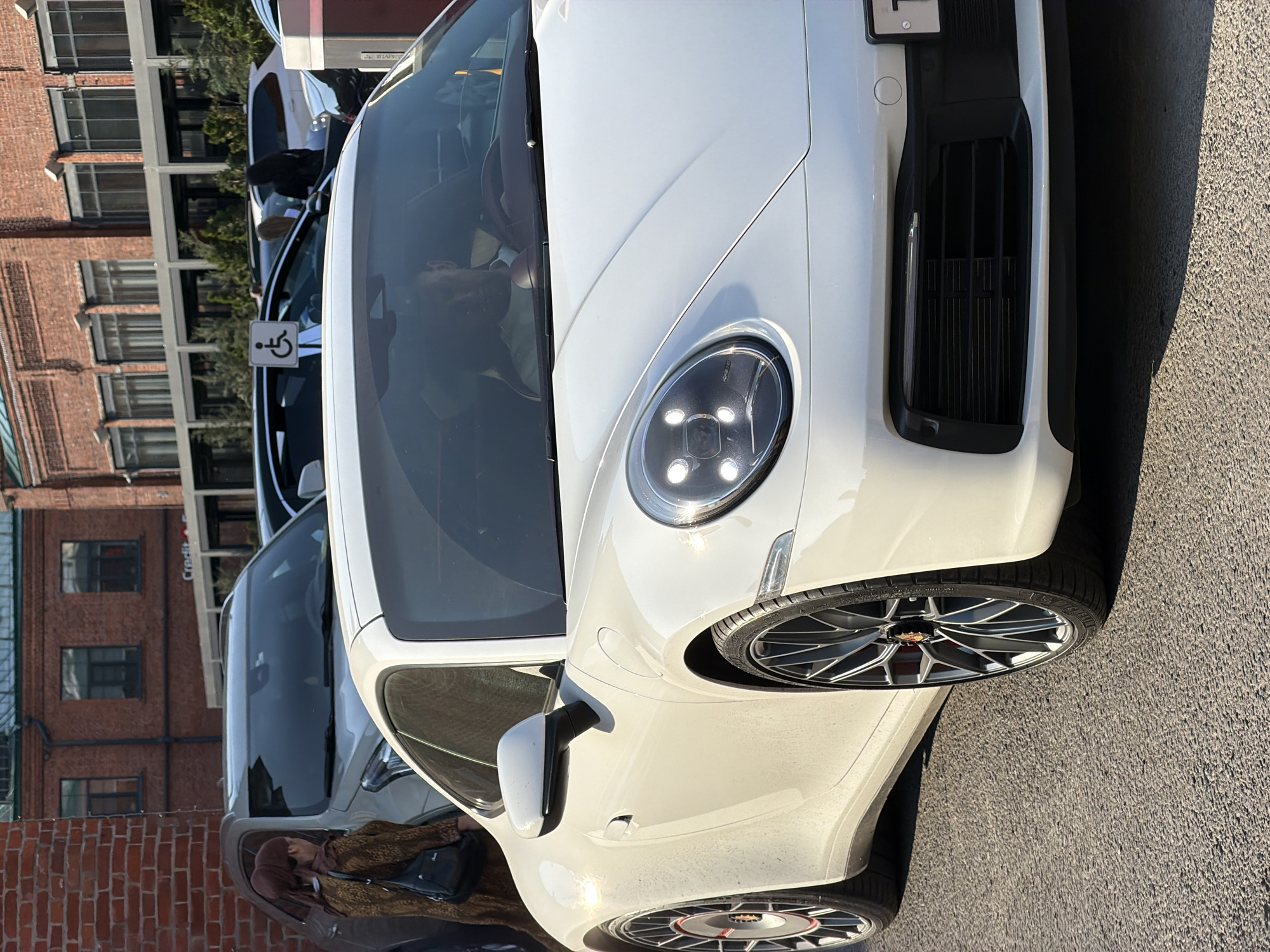 The image shows a white Porsche parked on a street. In the background, there is a red brick building with many windows, suggestive of an urban setting. The car is facing left and positioned next to another car. To the right of the car is a paved surface. Based on these observations, the location could be a street in Moscow, Russia, though specific landmarks are not visible in the image to confirm this definitively.