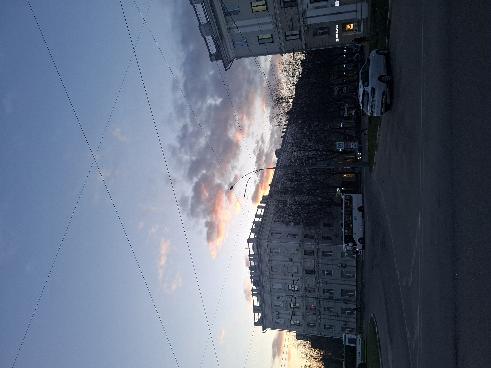 This image captures a city street scene during what appears to be dusk or dawn. The sky, occupying the upper portion, is a clear blue fading into lighter hues towards the horizon, with scattered clouds reflecting warm orange and pink light, indicative of sunrise or sunset. Overhead, multiple thin power lines or wires crisscross the sky.
On the left side of the street, several multi-story buildings with numerous windows line the road. One prominent building displays a sign with Cyrillic text, reading "ТРАПЕЗАРИЯ" (Trapezariya, meaning Refectory or Dining Room) with "СКИДКИ 60%" (Skidki 60%, meaning Discounts 60%) underneath. Bare trees, likely deciduous, stand between the buildings and the street, suggesting late autumn, winter, or early spring.
The street itself is dark asphalt, with a curving section in the foreground. On the right side of the road, a white car (license plate "В085АС 150" is visible, suggesting Moscow Oblast, Russia) is parked. Further down the street on the right, a white bus is partially visible, with "ЛОРЕ" (Lore) text on its side. Another green and white bus, displaying "БИОГАЗ ПРО" (Biogaz Pro), is partially seen at the bottom right. A street light with a curved pole extends into the frame from the right, its lamp visible. The overall mood is quiet and transitional, characteristic of a city winding down or waking up.