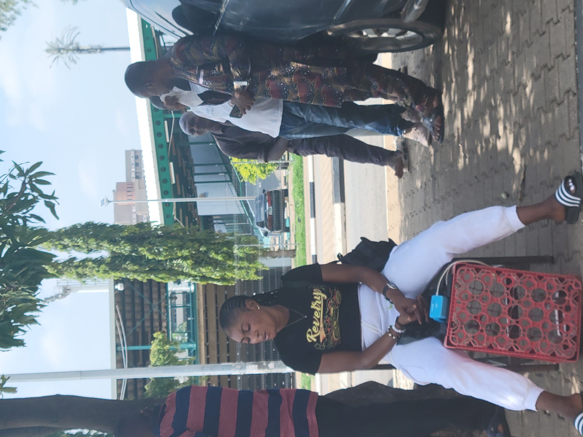 This image captures an outdoor street scene in Abuja, Nigeria, during bright daylight hours. The picture is rotated 90 degrees clockwise.
In the foreground, a woman is seated, looking down at her hands. She is wearing a black t-shirt with the word "Revelry" printed in gold, stylized font, and white pants, along with black and white striped slide sandals. Next to her is a red plastic crate, and a small blue electronic device (possibly a portable speaker or power bank) rests near her lap. She also has a black bag draped over her.
Behind the seated woman, a black car is partially visible. Several men are standing near the car. One man wears a patterned long-sleeved shirt and dark pants, while another wears a white t-shirt, blue jeans, and light-colored sandals. A third man, further back, is in a brown shirt and pants. The ground is paved with grey, patterned tiles, typical of a sidewalk.
In the background, there are green trees and foliage, various buildings, and a distinct tall communication mast or tower against a clear, bright sky. The scene appears to be a casual moment in an urban environment, with people seemingly waiting or resting under sunny weather conditions.
