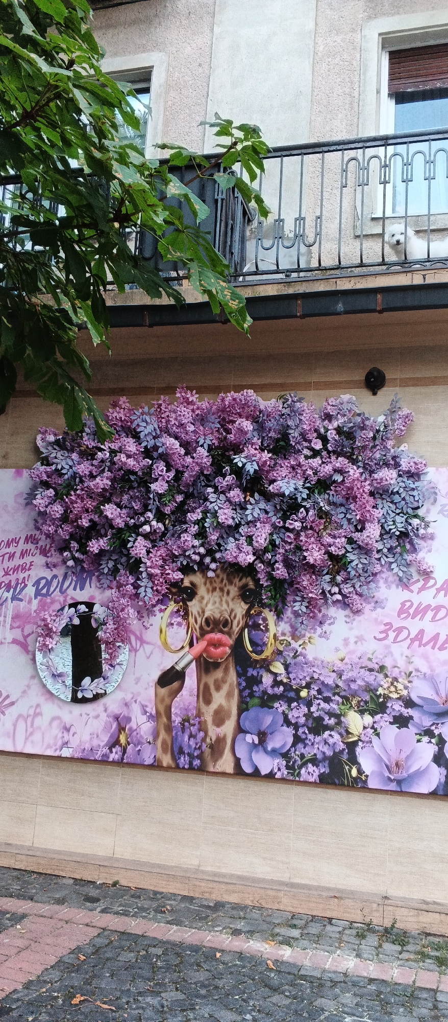 The image shows a mural of a giraffe wearing large hoop earrings and applying lipstick to its lips, with its head adorned by a large bloom of purple and pink flowers. The mural is on the exterior wall of a building, with a balcony railing visible above. Green leaves from a tree hang into the upper left portion of the frame. The mural is situated on a beige tiled wall above a cobblestone sidewalk. The text "ROOM" is visible on the mural in graffiti style. There is a small circular mirror with a floral frame attached to the left side of the mural. The image is taken outdoors during the day. The scene is located in Mukachevo, Ukraine.