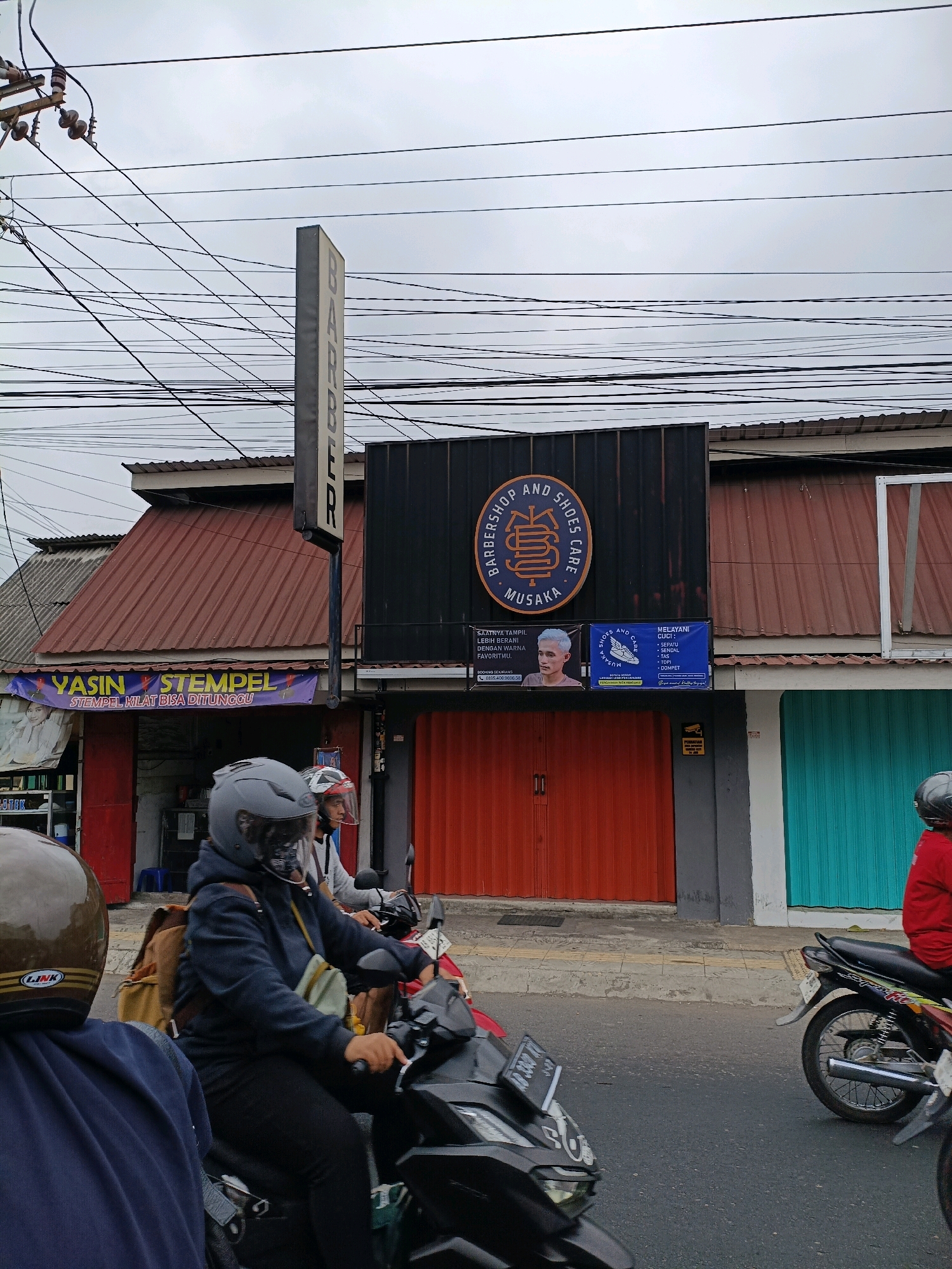 An exterior, eye-level, full shot captures a street scene in Yogyakarta, Indonesia, with motorcycles and a barber shop. The sky is overcast and gray, and a dense network of electrical wires is visible overhead. A tall, vertical sign with the word "BARBER" in gray capital letters is positioned to the left of the frame. Below the barber sign, a dark rectangular facade features a circular logo and the text "BARBERSHOP AND SHOES CARE MUSAKA." Smaller signs are mounted beneath this main facade. To the left, a red-fronted shop displays a banner that reads "YASIN STEMPEL" with "STEMPEL KILAT BISA DITUNGGU" below it, and a portrait of a person. To the right of the barber shop are two shuttered storefronts, one orange and one turquoise.
In the foreground, three people on motorcycles are moving along the street. The closest motorcycle on the left has its front wheel and license plate partially visible. The rider of the central motorcycle is wearing a gray helmet and a dark blue hooded jacket with a tan backpack. To their right, another person on a motorcycle wears a red jacket. The street is paved, and a sidewalk runs alongside the shops. The overall impression is of a typical urban street in Indonesia.