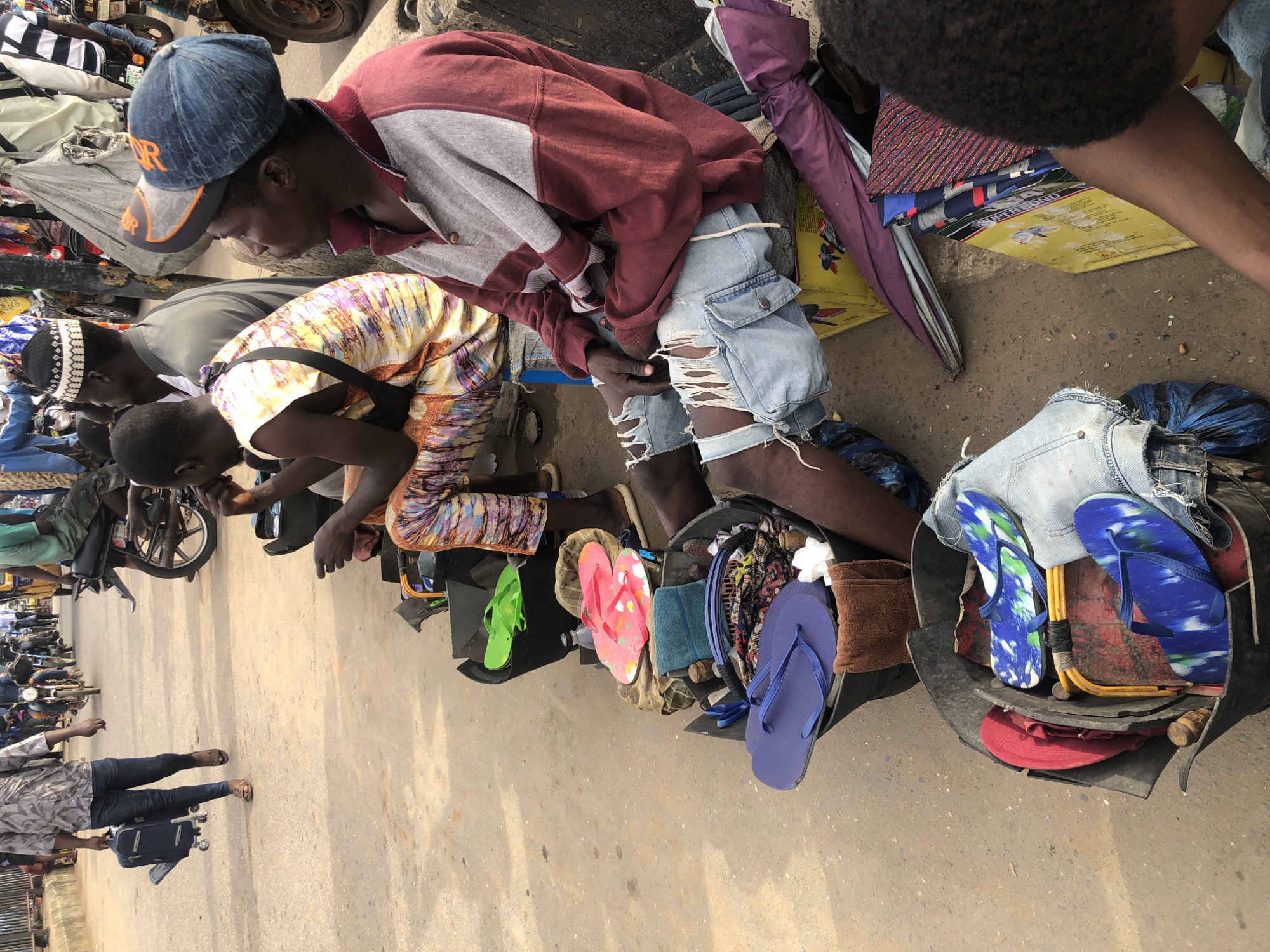 The image depicts an outdoor scene in a bustling urban environment, likely a market or roadside stall, in Ikeja, Nigeria. The focus is on a group of men, some seated and others partially visible. In the foreground, a man wearing a maroon and gray long-sleeved shirt and distressed denim shorts is seated, looking to his right. Next to him, a bucket overflows with various items, including blue flip-flops and what appear to be patterned fabric pieces. Another bucket to the left contains more flip-flops, including bright pink ones and green ones. In the middle ground, another man in a vibrantly patterned sleeveless shirt is seated, also looking to his right. Behind them, several individuals are seen walking or standing, some with motorcycles nearby. One person in the background is seen pulling a rolling suitcase. The ground is paved with concrete or asphalt, and there are other assorted objects and debris scattered around, suggesting a busy and perhaps somewhat unkempt area. The overall impression is one of commercial activity and daily life unfolding on a street in Nigeria.