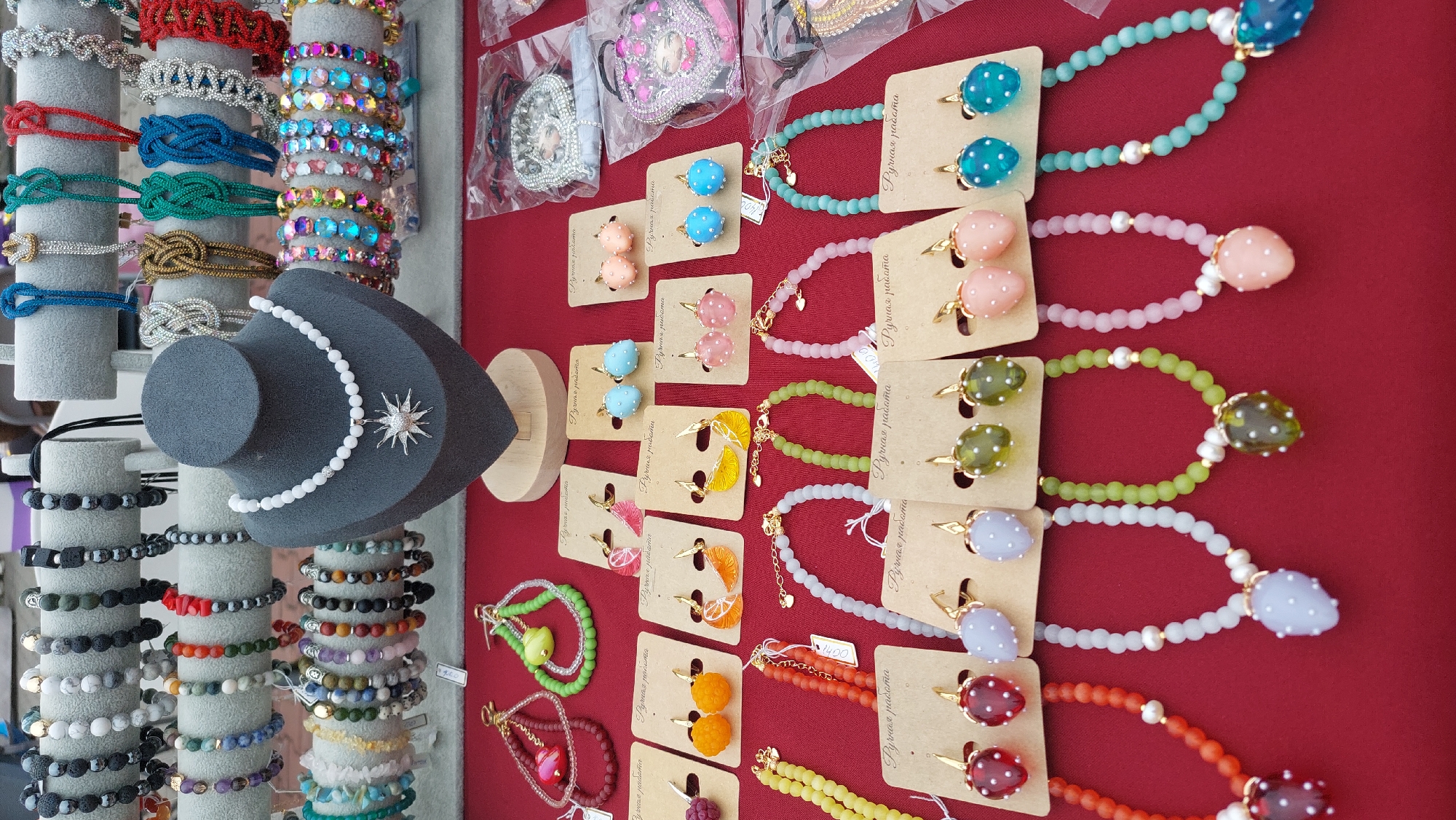 This image displays a stall at an outdoor market in Komsomolsk-on-Amur, Russia, featuring a variety of handmade jewelry. The stall has a red tablecloth displaying numerous bracelets and necklaces. In the background, more bracelets are arranged on gray stands. In the foreground, several pairs of earrings are presented on cardboard backing, with corresponding necklaces displayed below them. The jewelry includes items made of beads in various colors, shapes, and sizes, with some pieces featuring strawberry or fruit motifs.